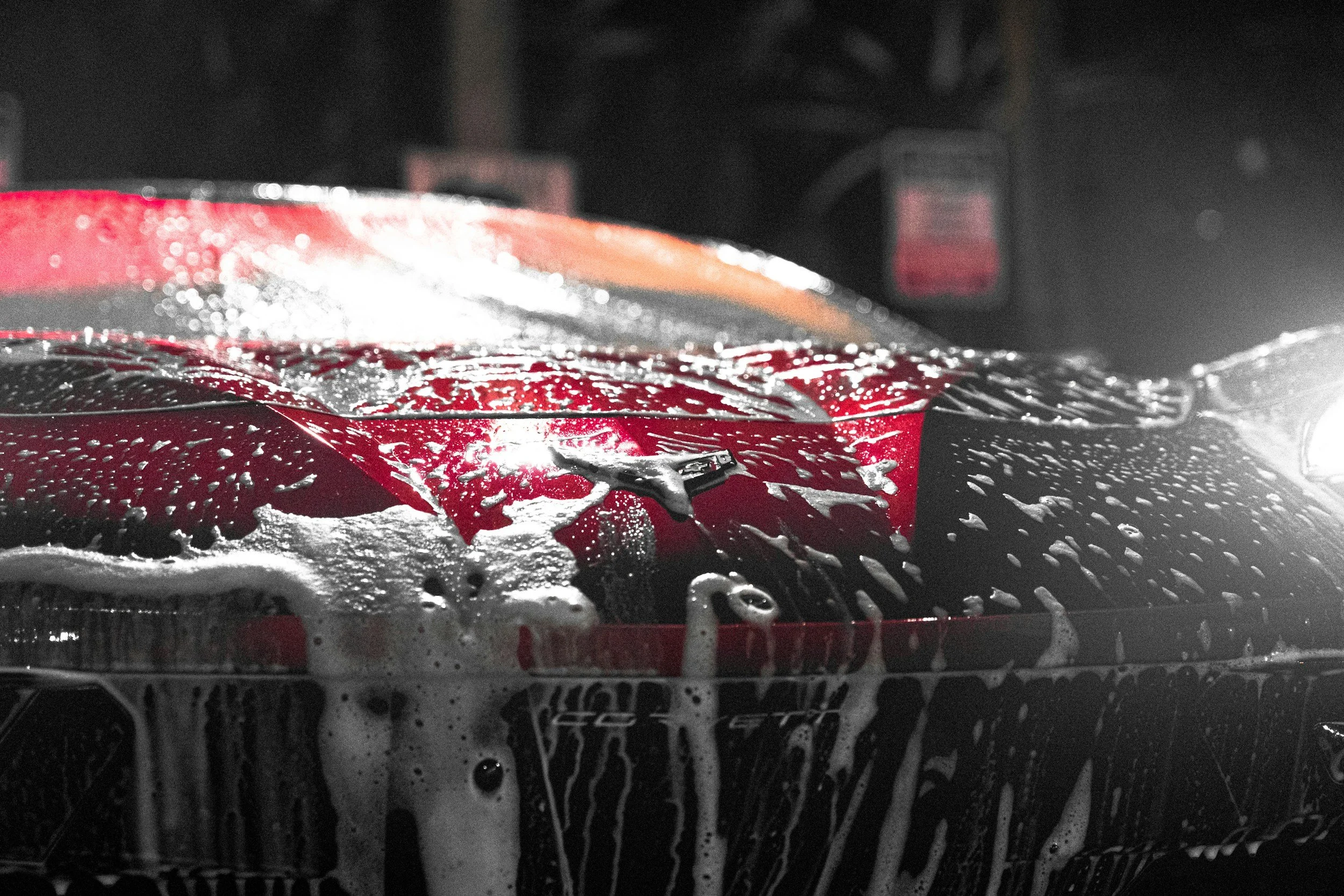 Close-up of a red car being washed with soap and water, foam and bubbles visible on the surface, at night.