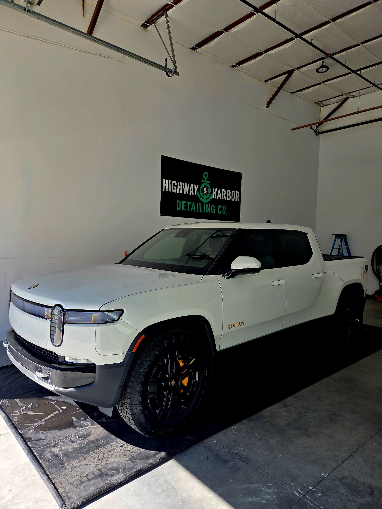 A white Rivian R1T electric pickup truck parked inside a garage with a high ceiling, with a sign on the wall that reads 'Highway & Harbor Detailing Co.'