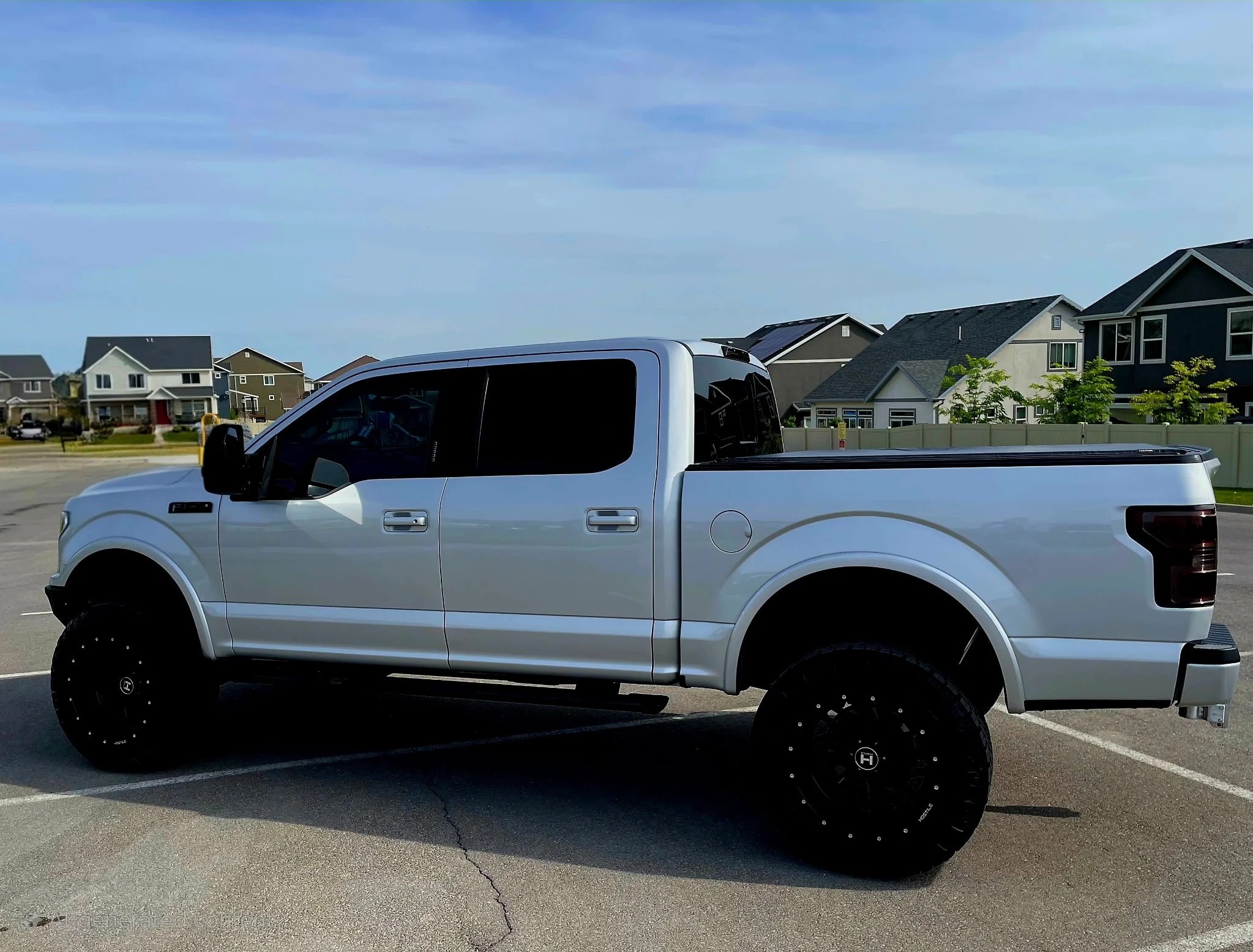 White pickup truck with black wheels parked in a lot near residential houses under a partly cloudy sky.