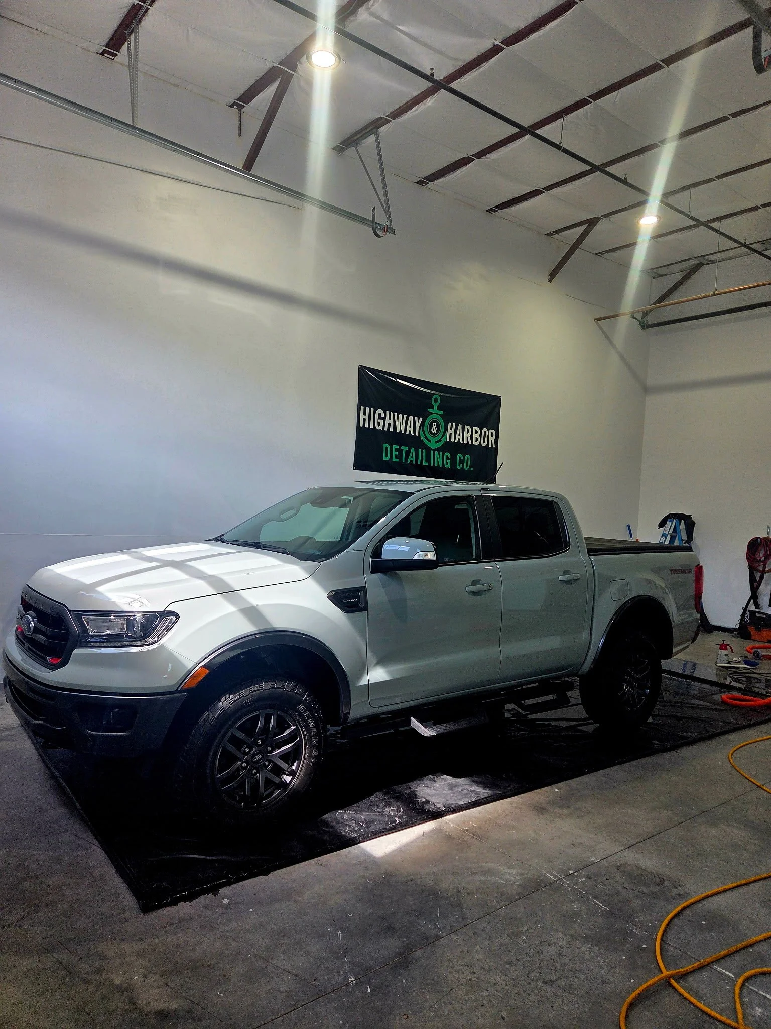 A white pickup truck inside a detailing garage with a black and green Highway & Harbor Detailing Co. banner on the wall