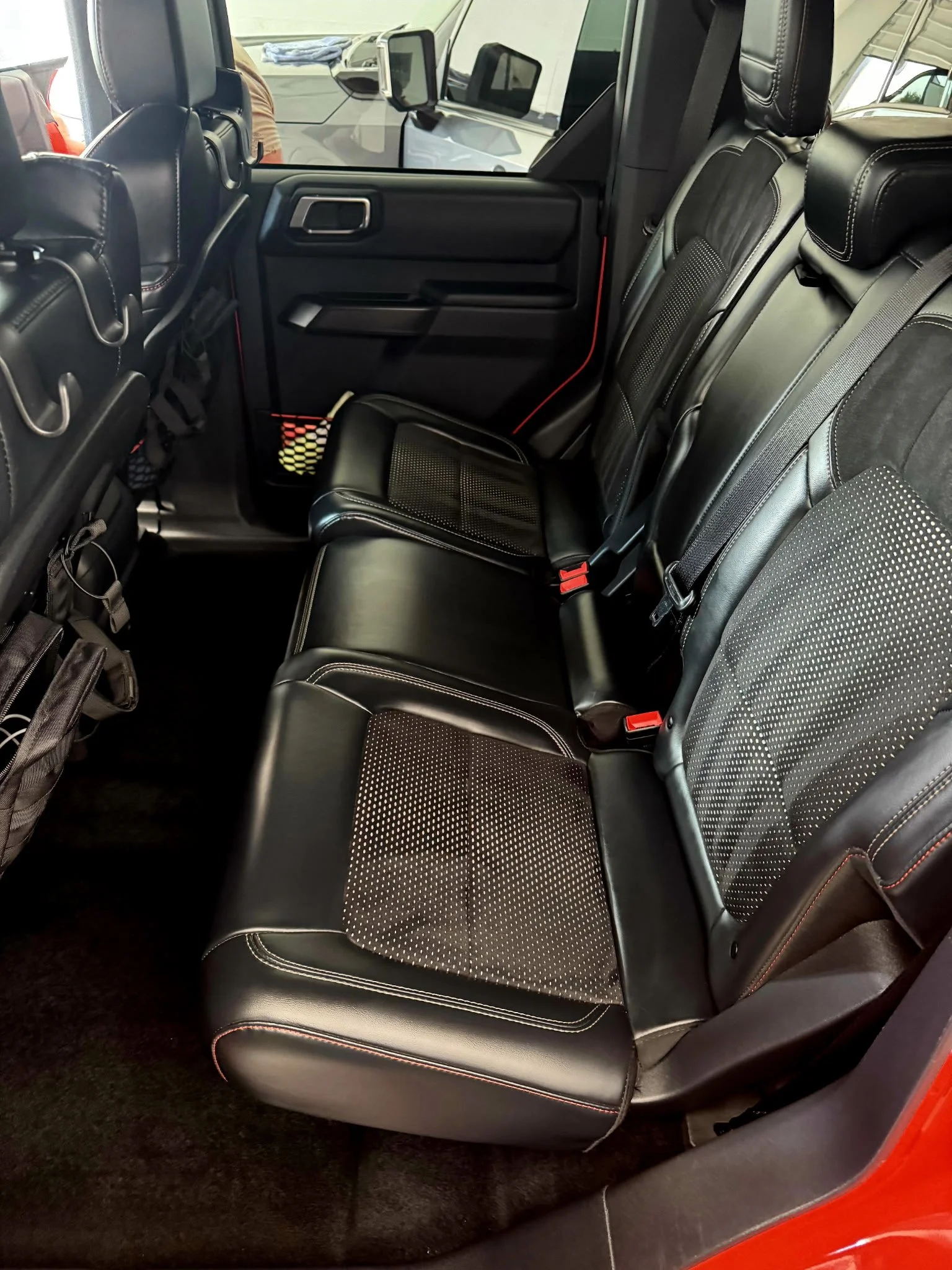 The back seat of a black vehicle with leather and fabric seats, featuring patterned fabric inserts, red stitching, and three seat belts.