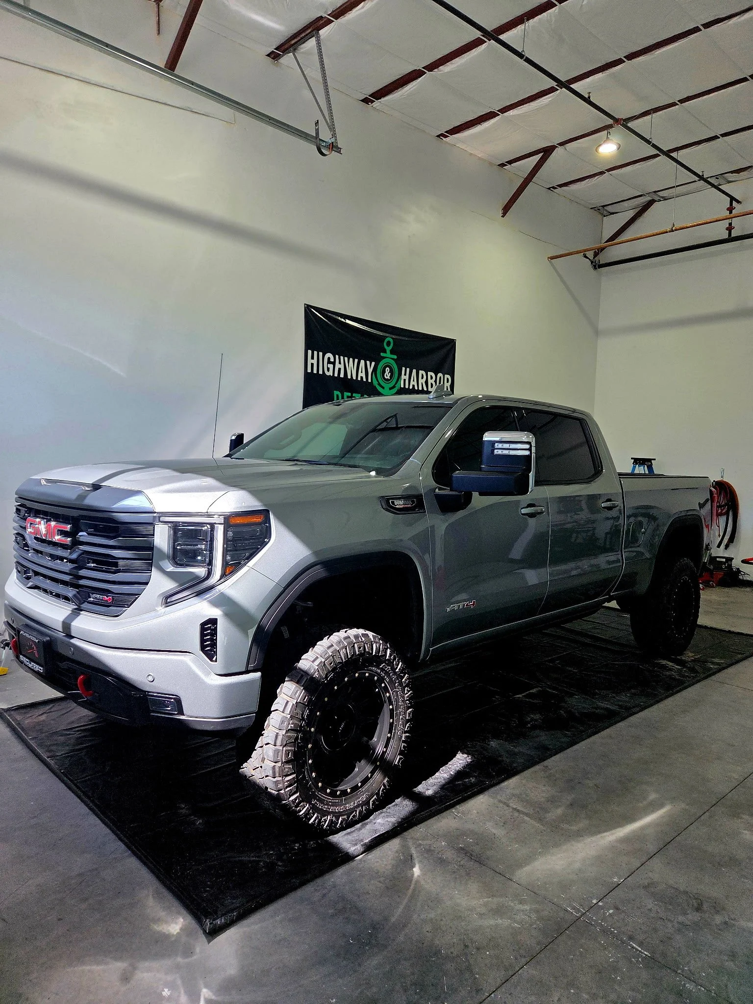 A silver GMC pickup truck with off-road tires parked inside a spacious indoor garage. The truck is on a black mat, and there is a highway and harbor decal banner on the wall behind it. The ceiling is high with metal beams and white insulation panels.