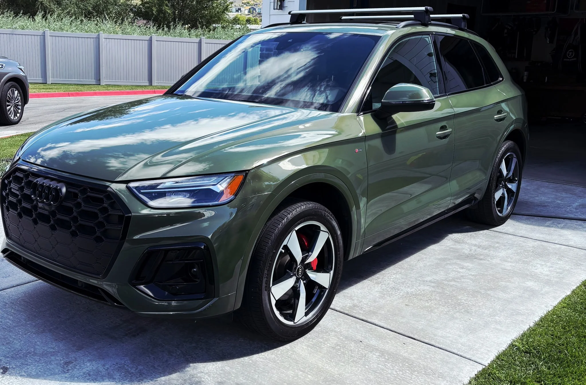 Green Audi SUV parked in driveway, reflecting clouds in the windshield, with a fence and other cars in the background.