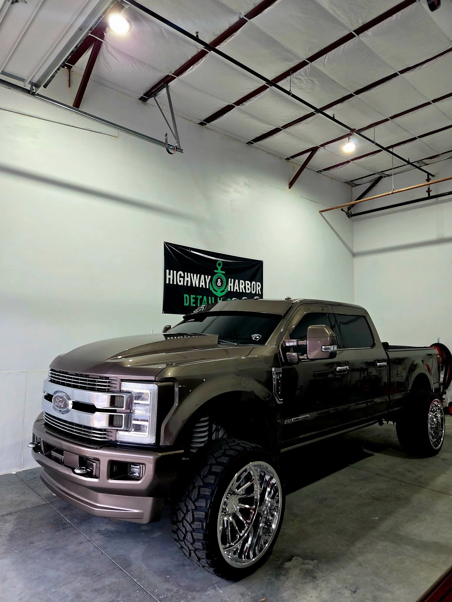 A modified Ford pickup truck with large chrome wheels and off-road tires is parked inside a garage or showroom. The truck has a dark gray or brown exterior and is positioned in front of a black sign that reads "Highway & Harbor Detailing." The garage