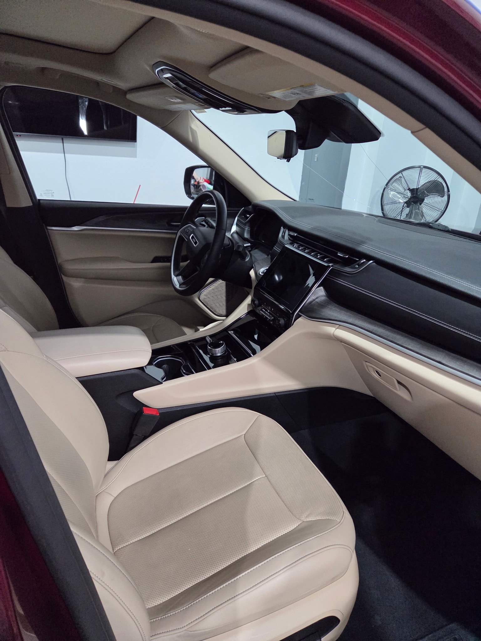 Interior of a modern vehicle with beige leather seats, a black dashboard, and a large central touch screen display, with a fan visible through the window.