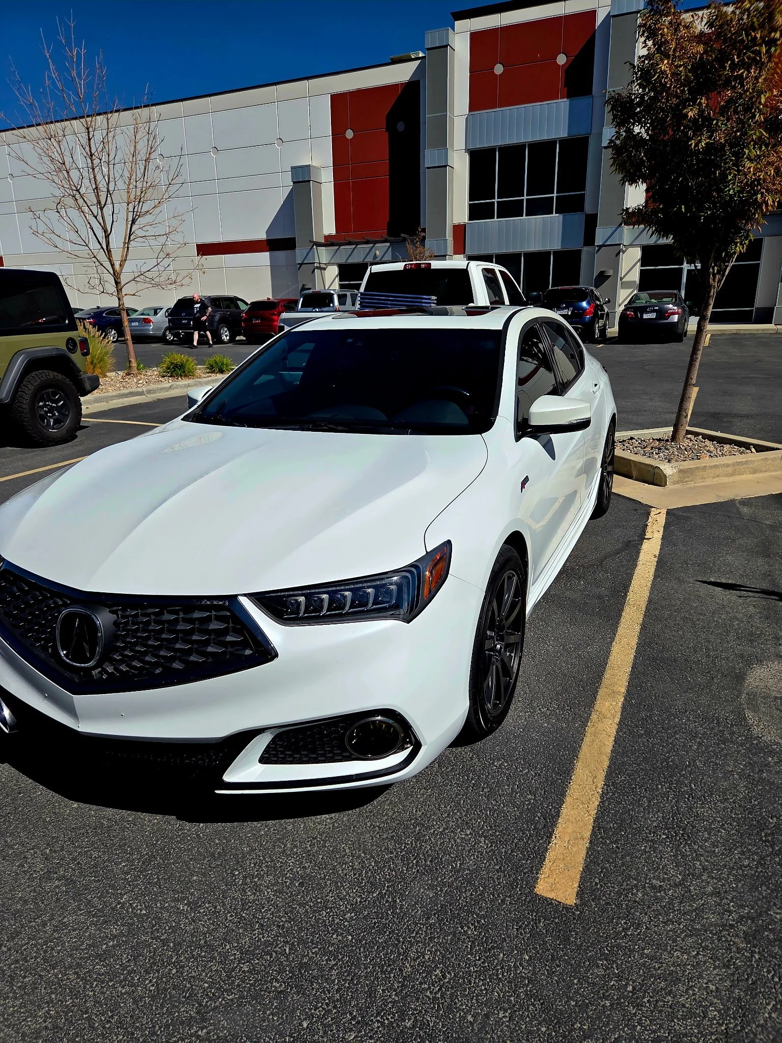 White sedan car parked in a parking lot next to trees, with a modern building in the background.
