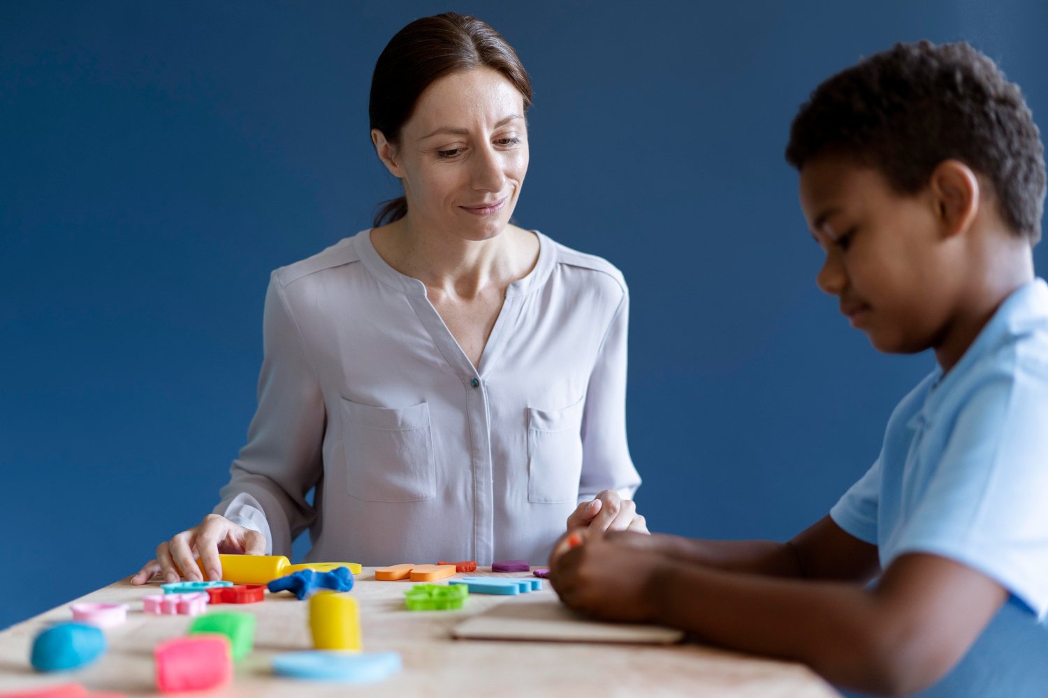 An RBT Therapist and a young boy playing with colorful alphabet and number foam puzzles on a wooden table against a blue wall.