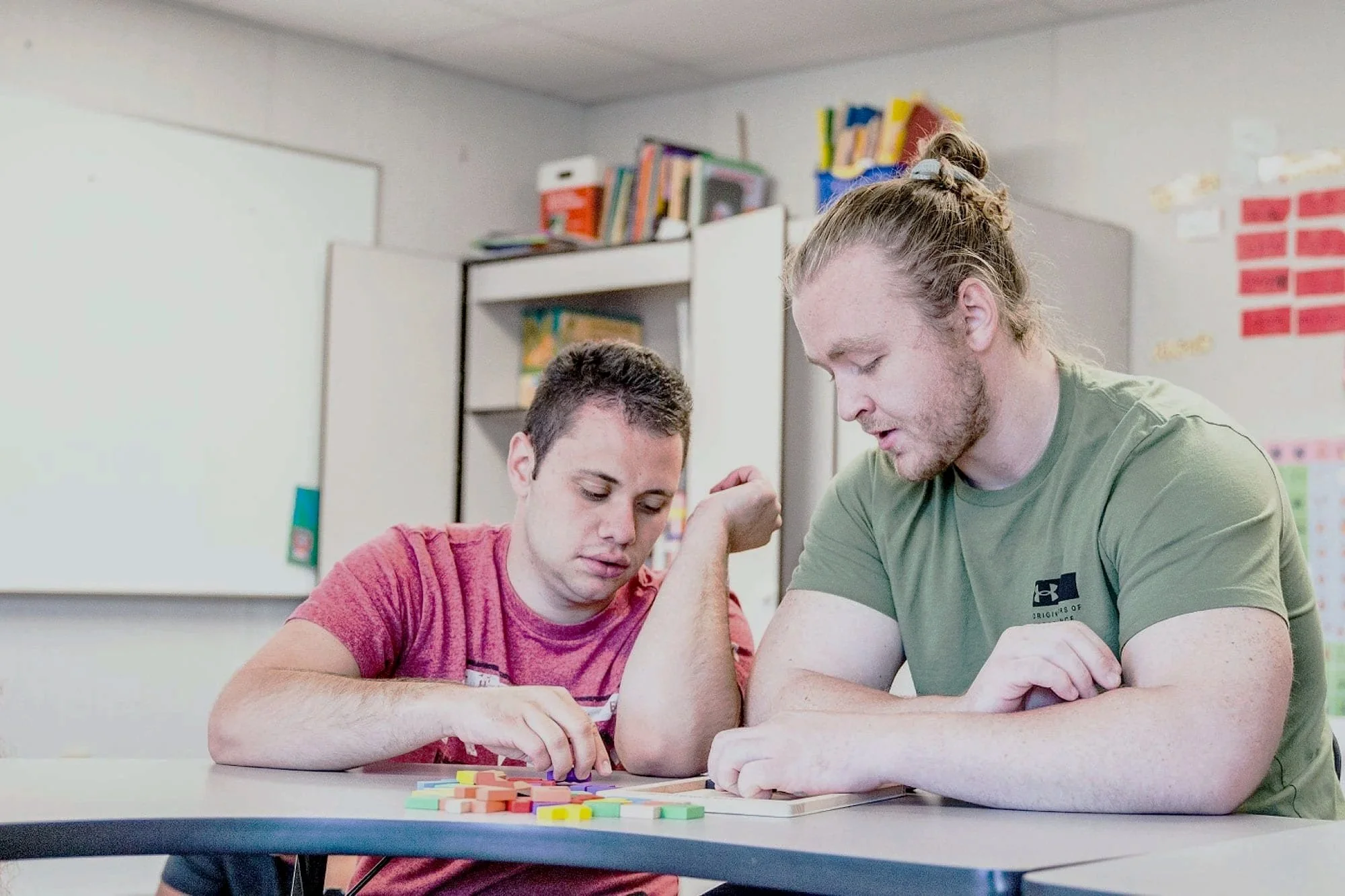 Two young men sitting at a table, playing a board game with colorful pieces in a classroom.