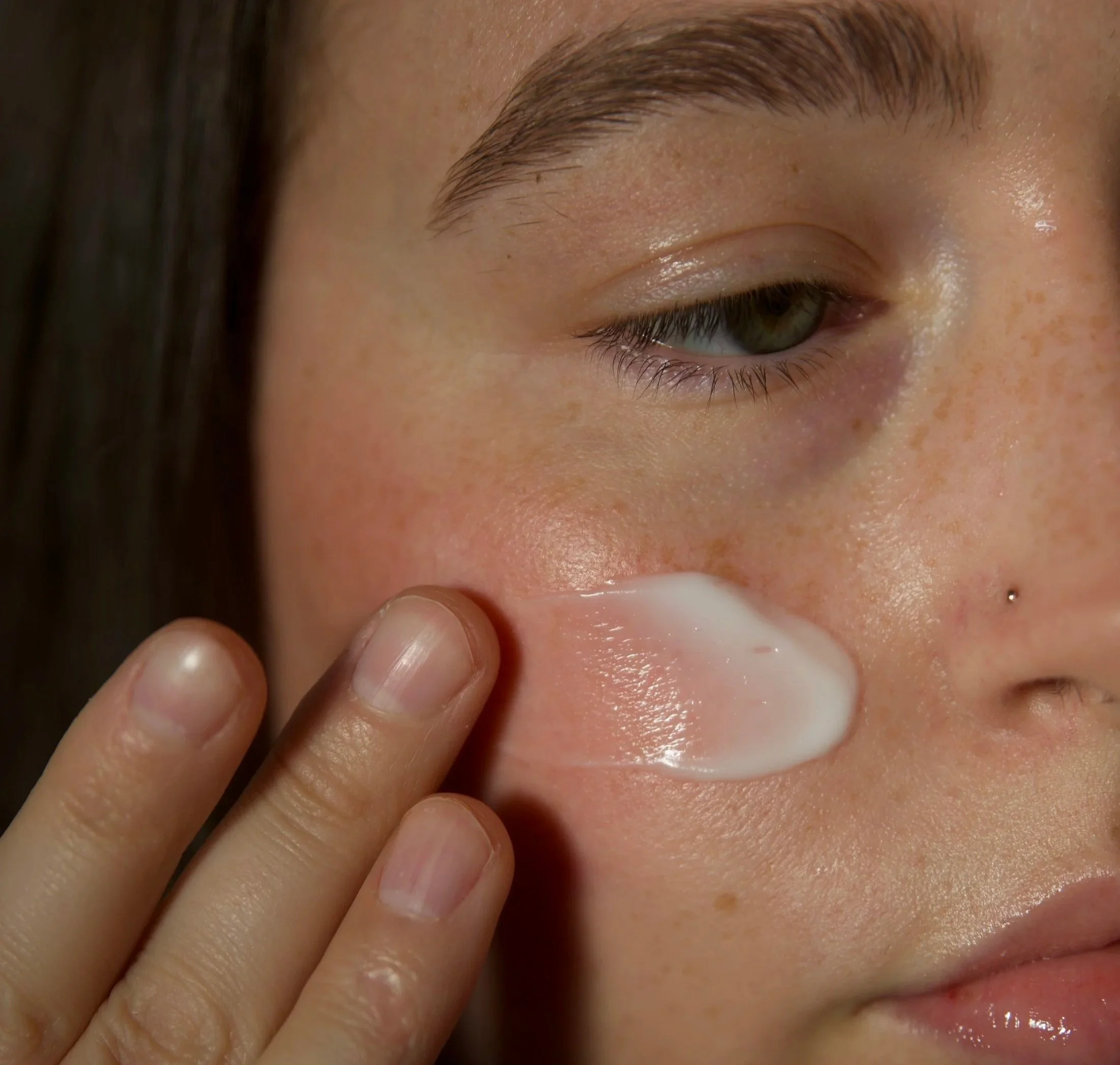A person with light skin and freckles applies white cream to the cheek with their index finger, with the face partially visible.