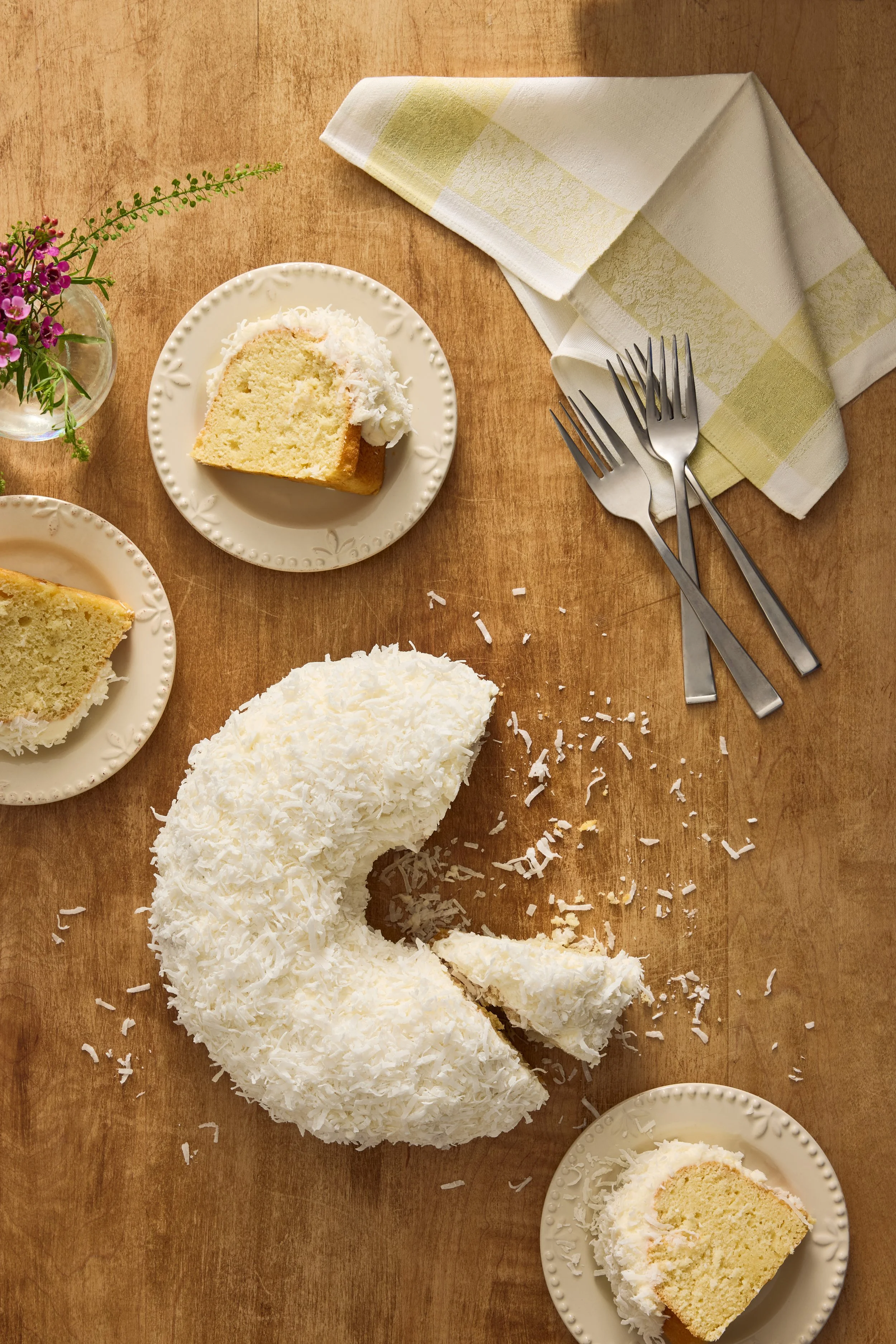 A coconut cake with shredded coconut on top and a missing slice on a wooden table, along with two slices of cake on small plates, a vase of flowers, and utensils.