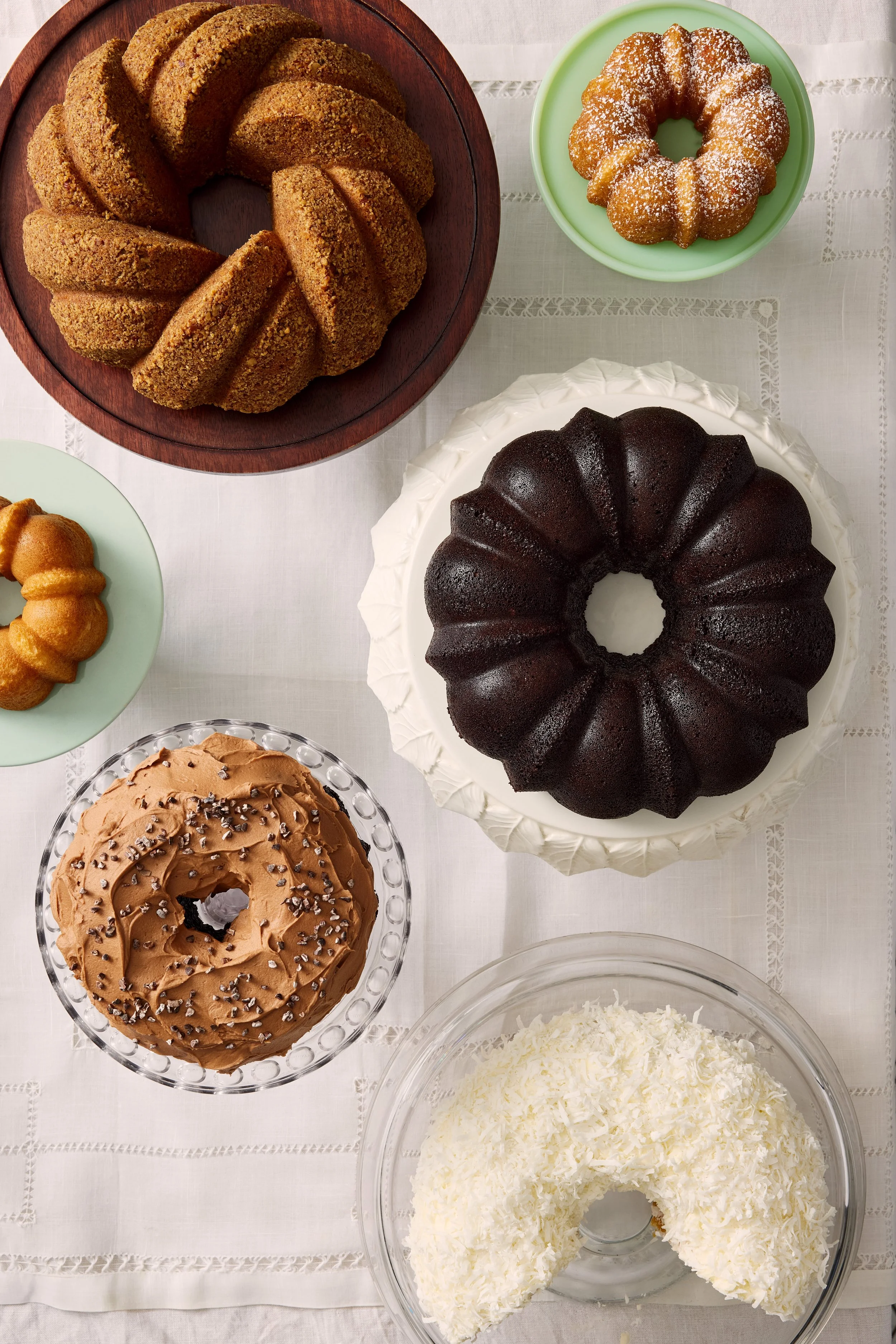 Assorted cakes and desserts on a table, including bundt cakes in various flavors, a cream-filled cake with coconut, and a chocolate cake, all on different plates.