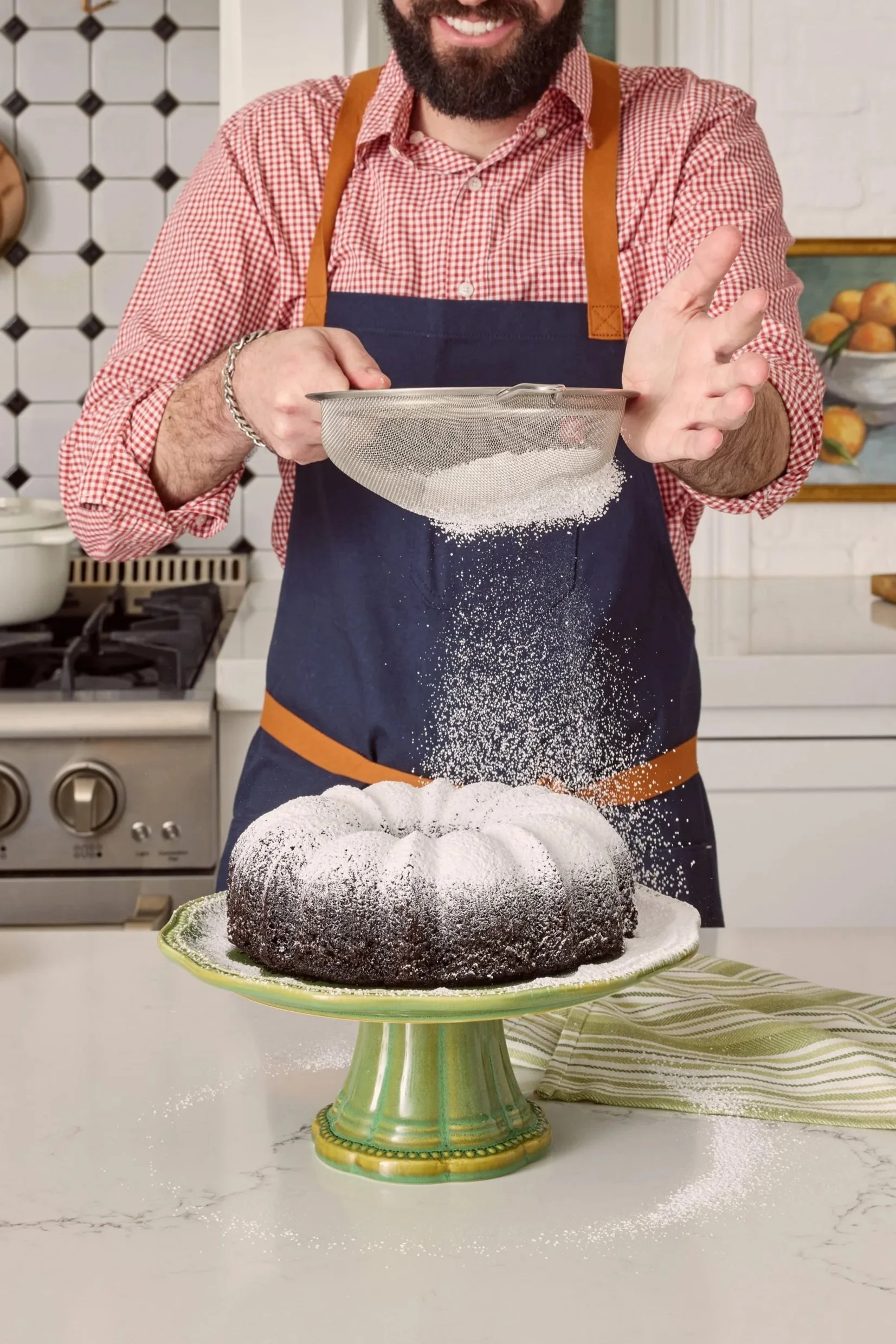 A man with a beard in a red checkered shirt and dark apron is dusting a chocolate Bundt cake with powdered sugar in a kitchen.