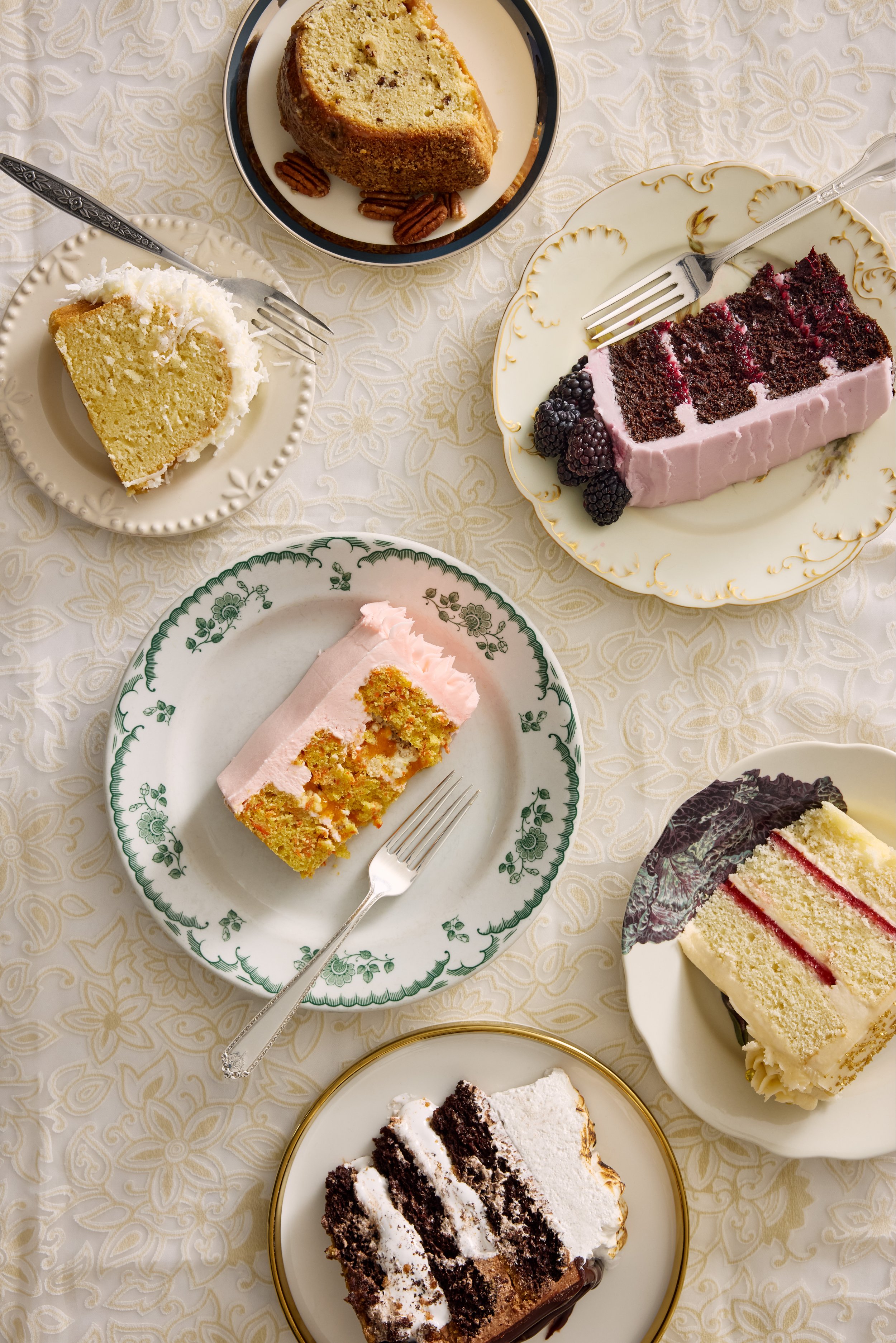 An assortment of six different slices of cake on elegant plates are arranged on a cream-colored tablecloth.