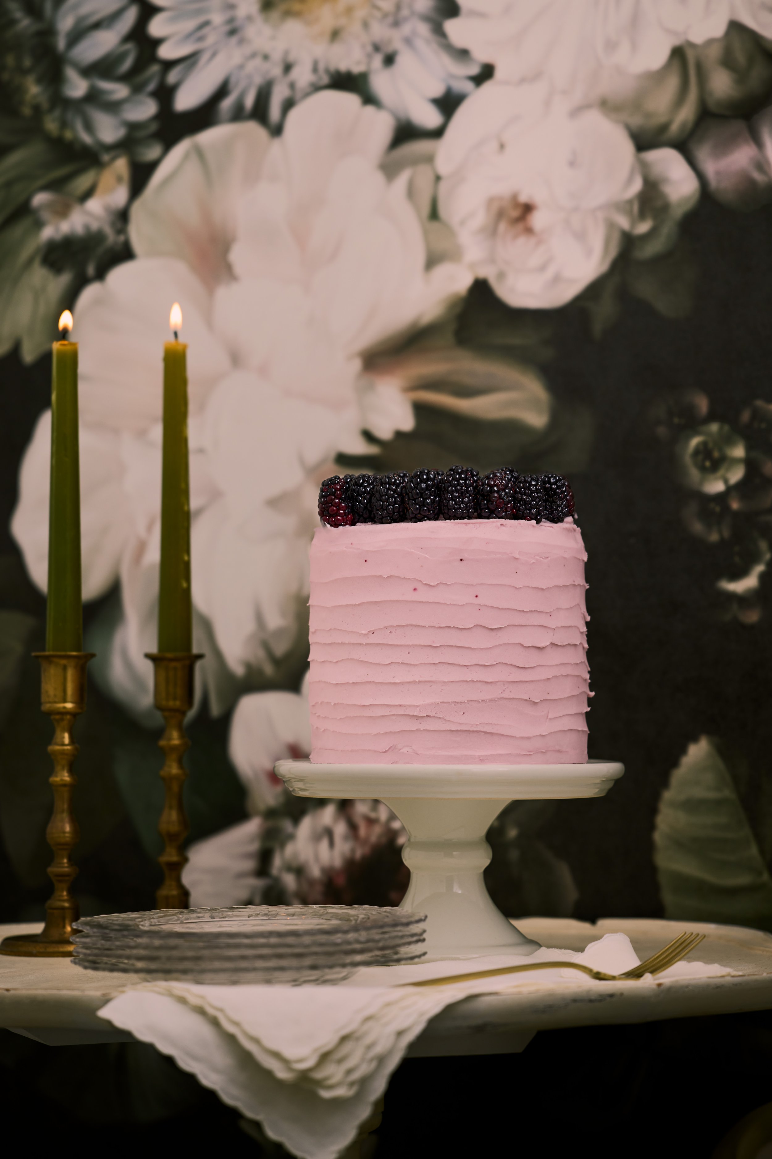 Pink frosted cake topped with blackberries, two lit green candles, and a stack of gray napkins on a white cake stand with a background of large white and green flowers.