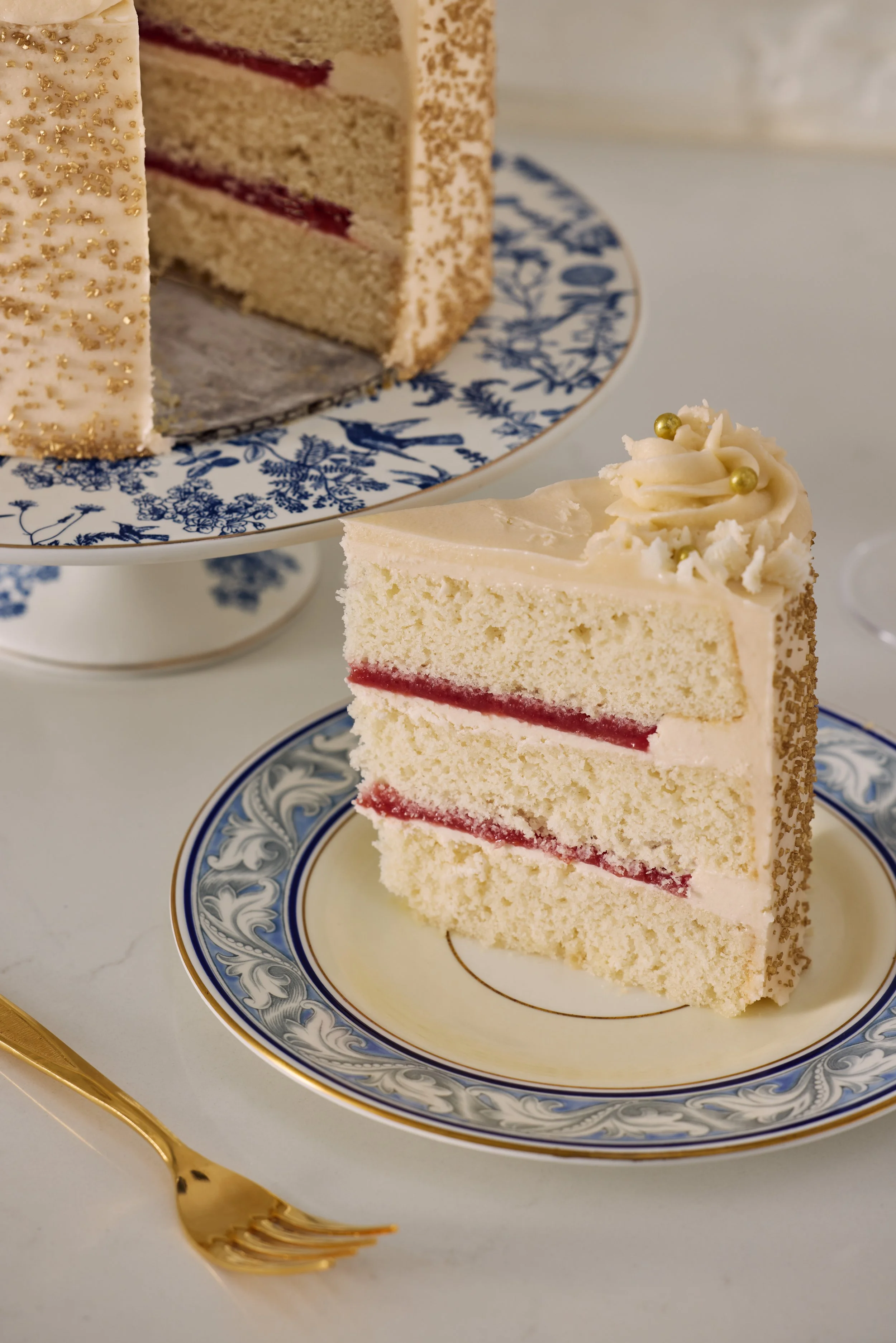 A slice of layered vanilla cake with red jam and cream filling on a decorative plate, with a larger whole cake in the background on a cake stand.