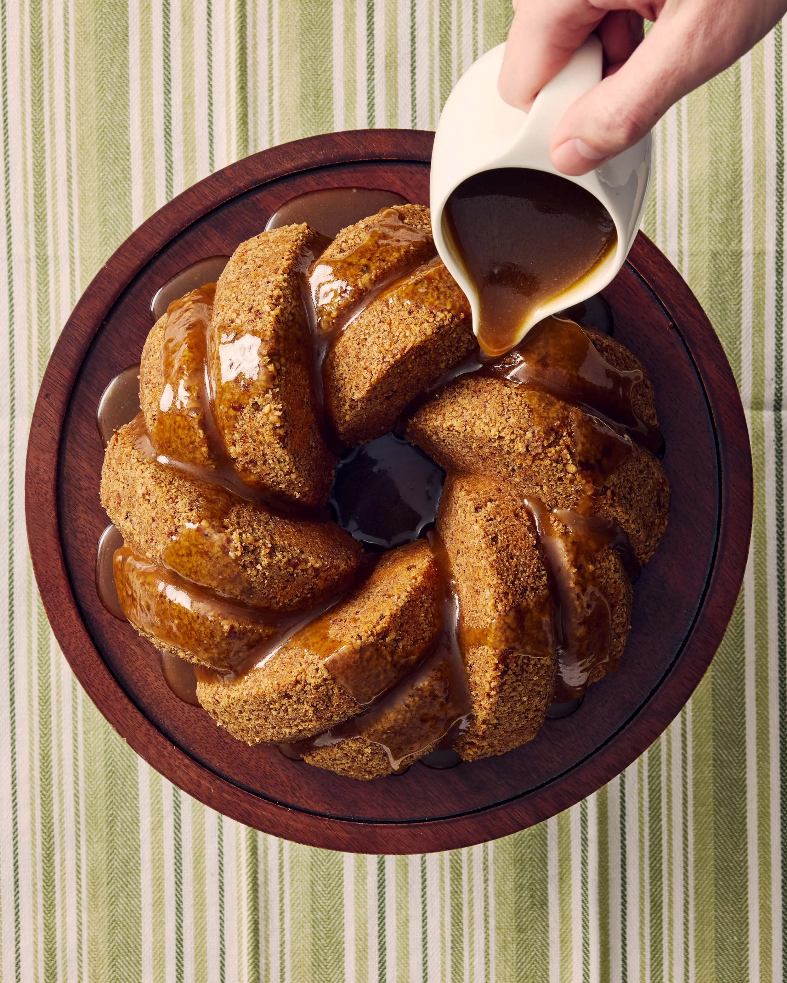 A Bundt cake with a graham cracker crust, topped with caramel sauce, sitting in a wooden serving dish on a green striped tablecloth. Someone is pouring caramel sauce over the cake.