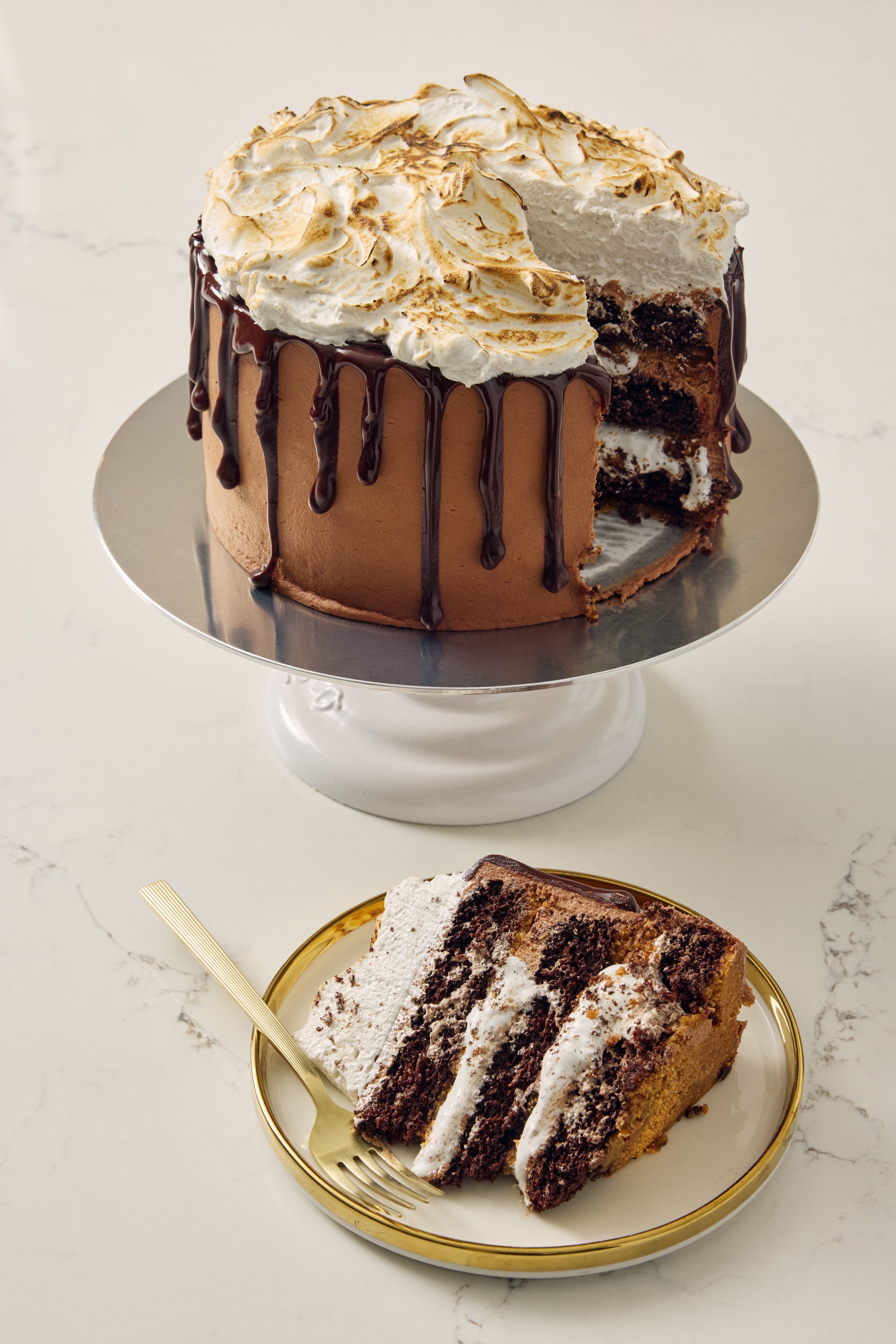 A chocolate layer cake with whipped topping and chocolate drizzle on a silver cake stand, and a slice of similar layered cake on a small gold-rimmed plate with a gold fork.