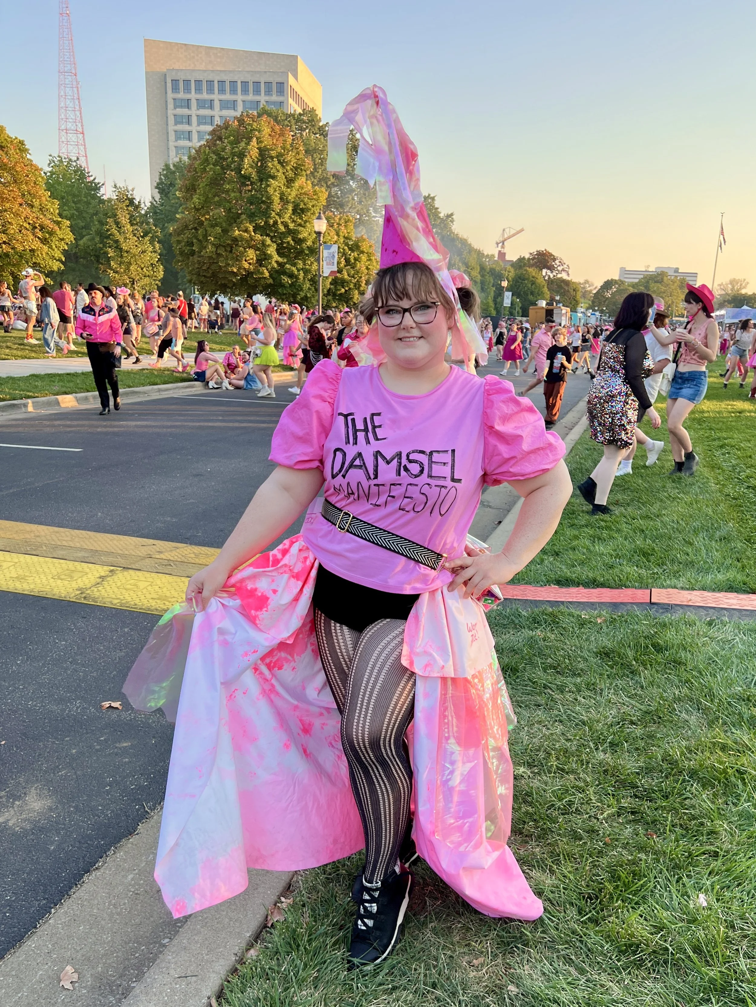 A young girl in a pink T-shirt with the words 'The Damsel Santa Fe' printed on it, standing outdoors during the daytime, surrounded by people at an event. She is wearing glasses, patterned tights, a pink satin sash, and a tall pink headpiece with colorful ribbons. There are trees, buildings, and a crowd in the background.