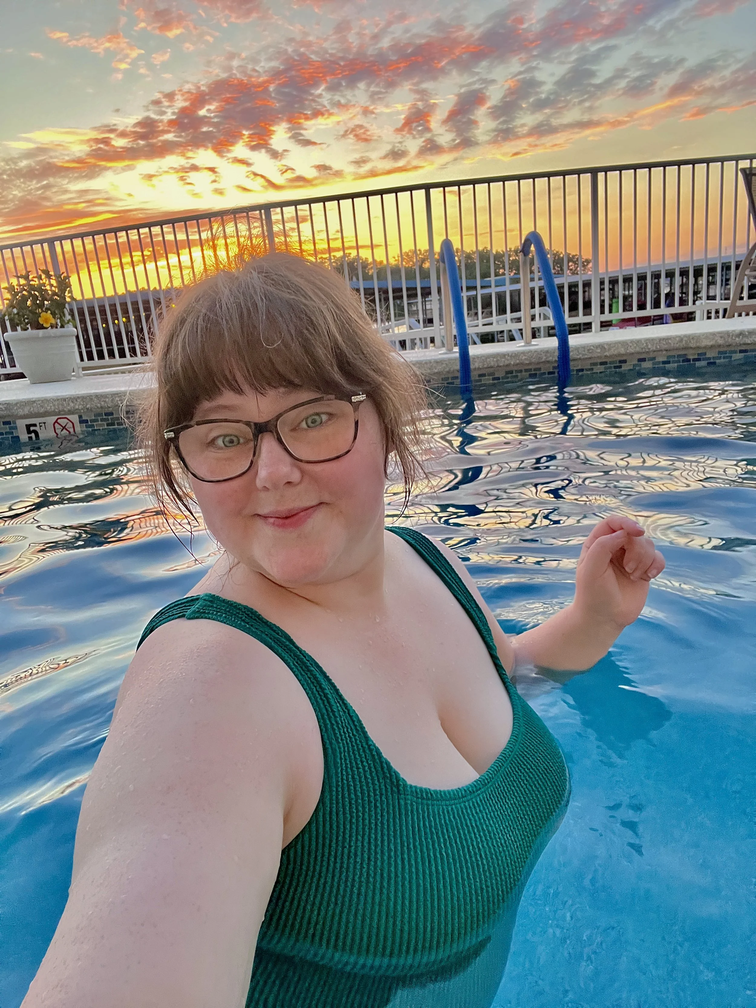 A woman wearing glasses in a green swimsuit taking a selfie in a swimming pool at sunset, with a sunset sky and a pool ladder in the background.