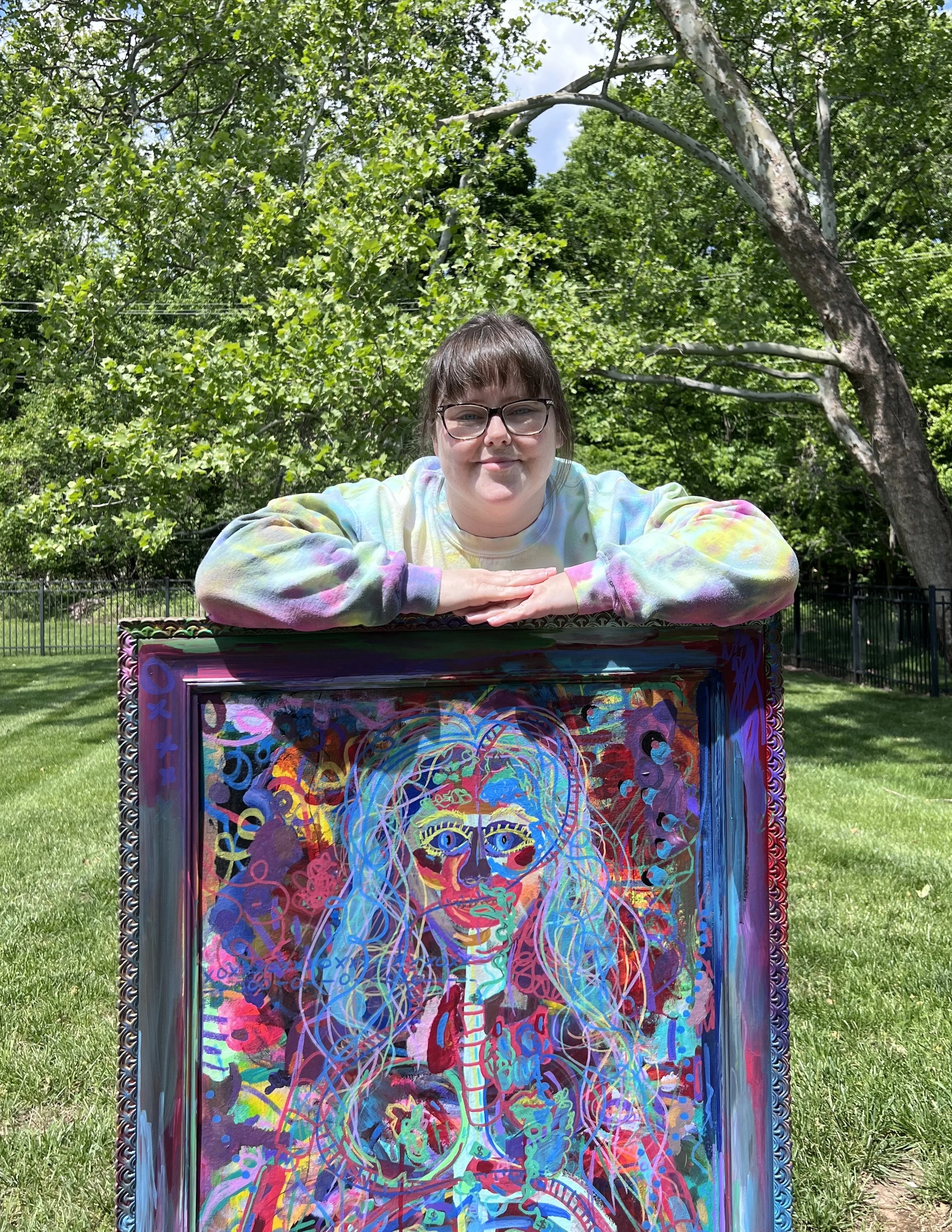 A woman with glasses and a tie-dye shirt is leaning on a colorful, abstract painting of a human skeleton with a cat face in a park on a sunny day.