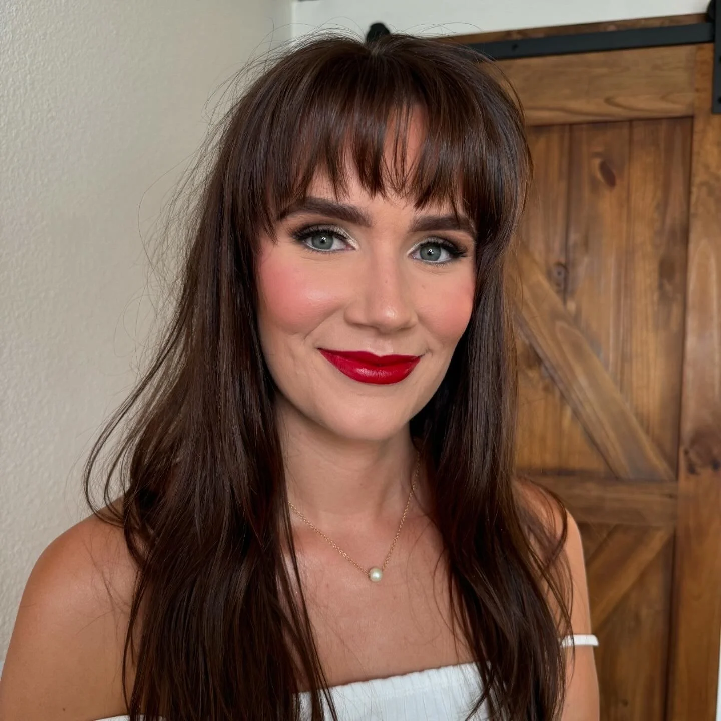 A woman with long brown hair, red lipstick, and blue eyes, wearing a pearl necklace and a white off-the-shoulder top, smiling indoors with a wooden door in the background.