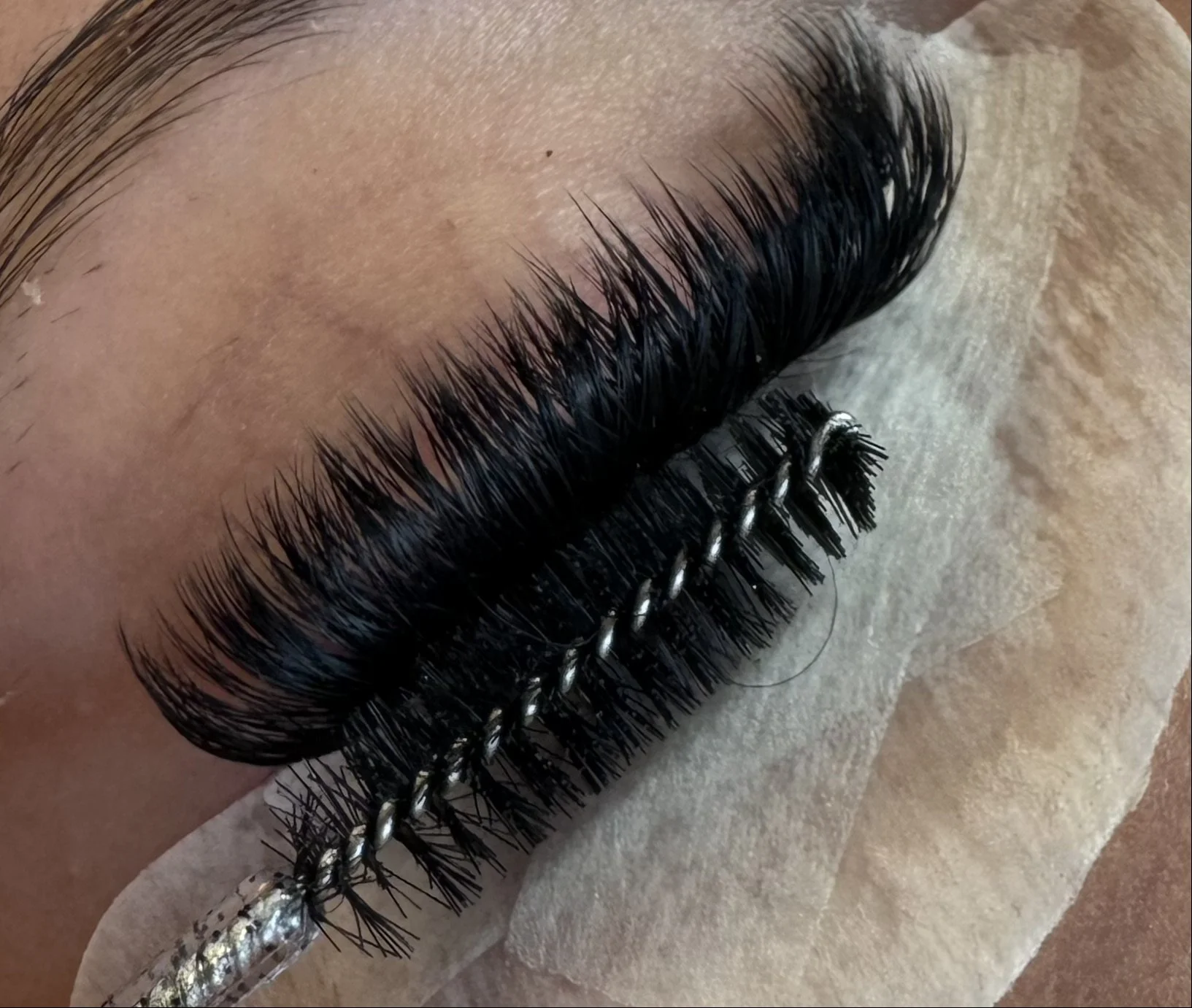 Close-up of a person's eye with long, thick, black eyelash extensions applied. An eyelash extension applicator is laid across the eye, with the person's eyelid resting on a strip of tape or pad.
