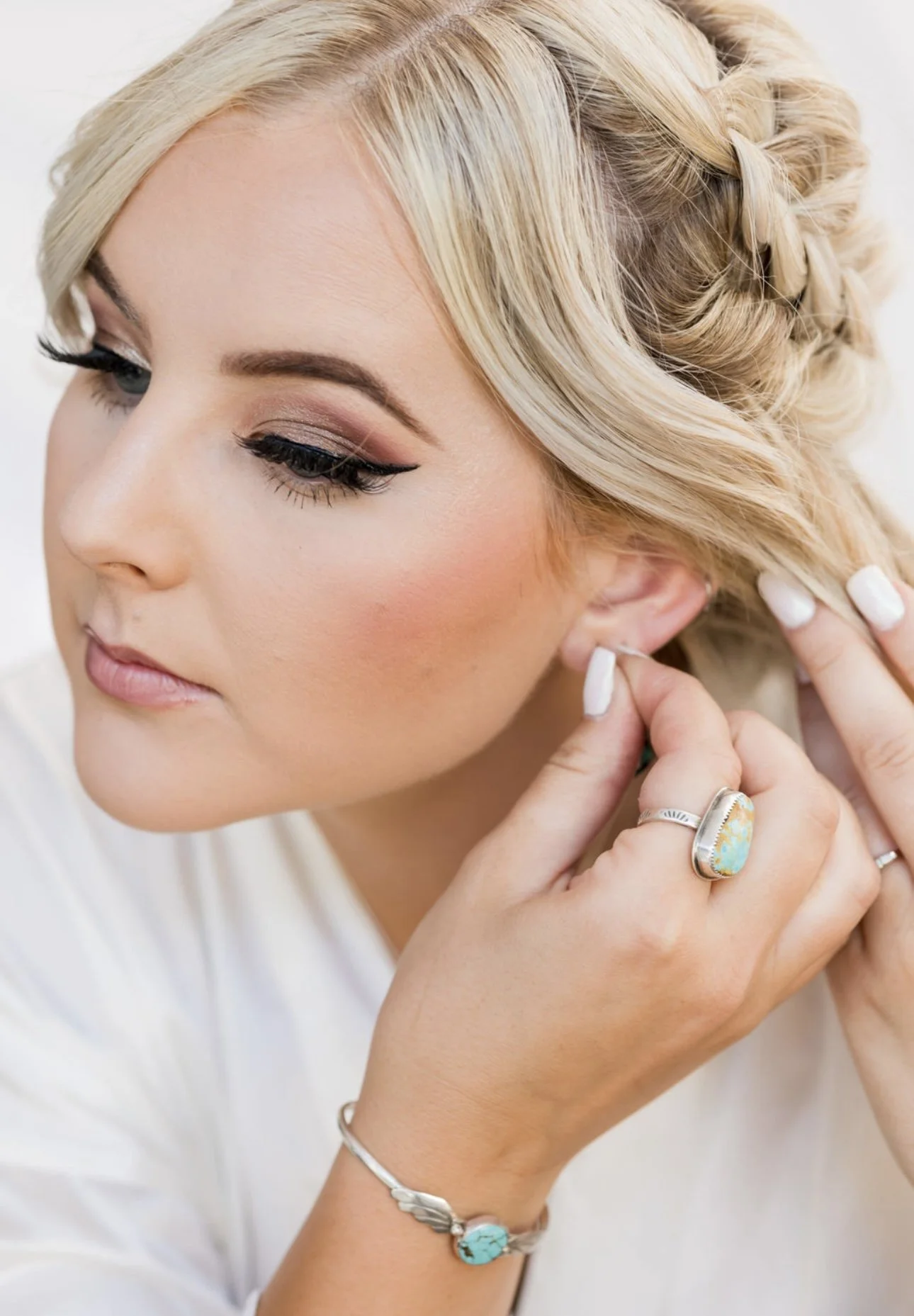 A woman with blonde hair styled in loose waves and a braid, wearing makeup, is putting on an earring.