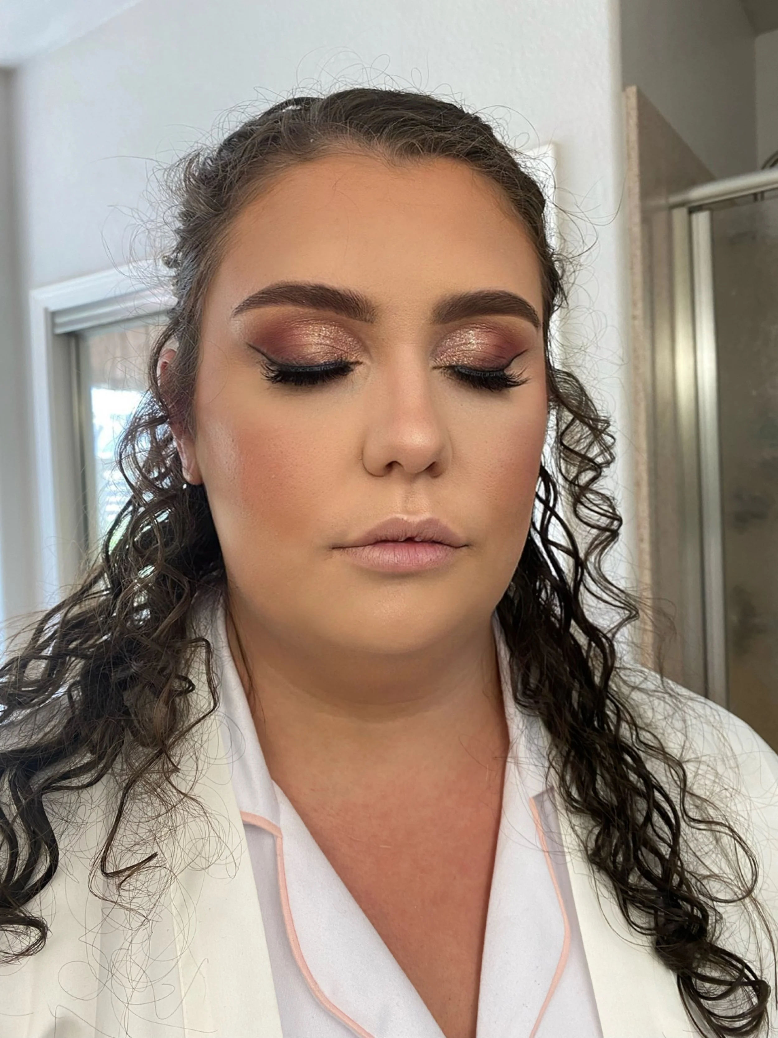 A woman with curly dark hair and makeup featuring shimmery eyeshadow, winged eyeliner, and nude lipstick, standing in a bathroom with closed eyes.
