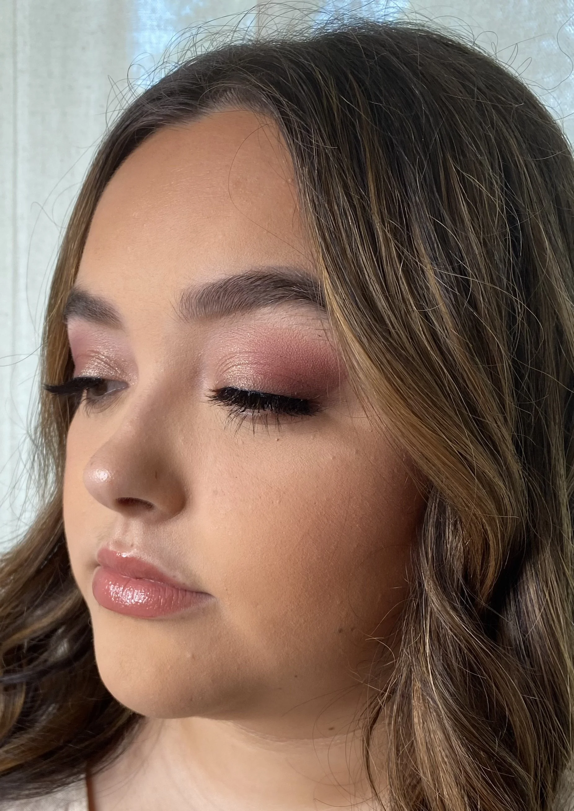 Close-up of a young woman's face with makeup, featuring glittery eyeshadow, long eyelashes, glossy lips, and wavy brown hair.