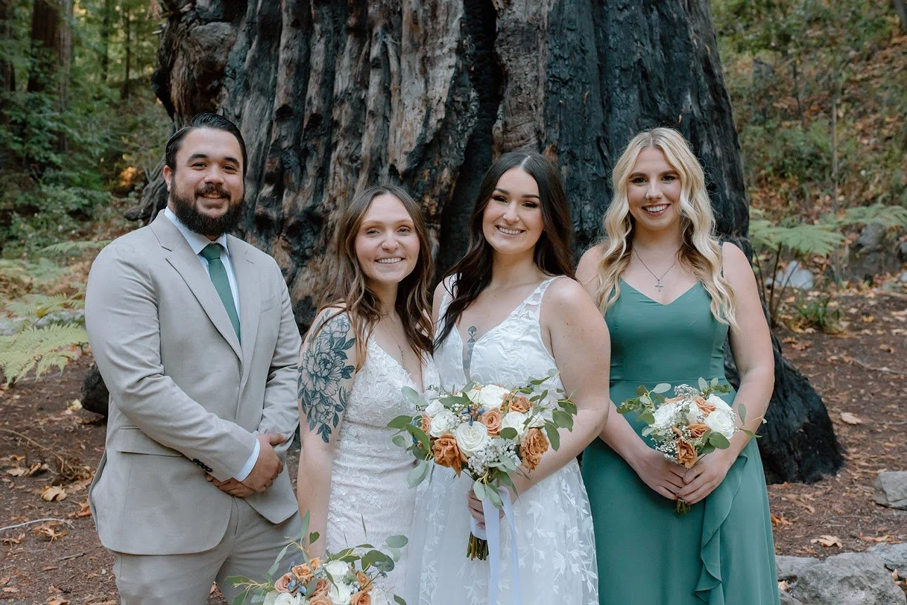 Group of four people dressed in wedding attire, standing outdoors in front of a large tree surrounded by nature.