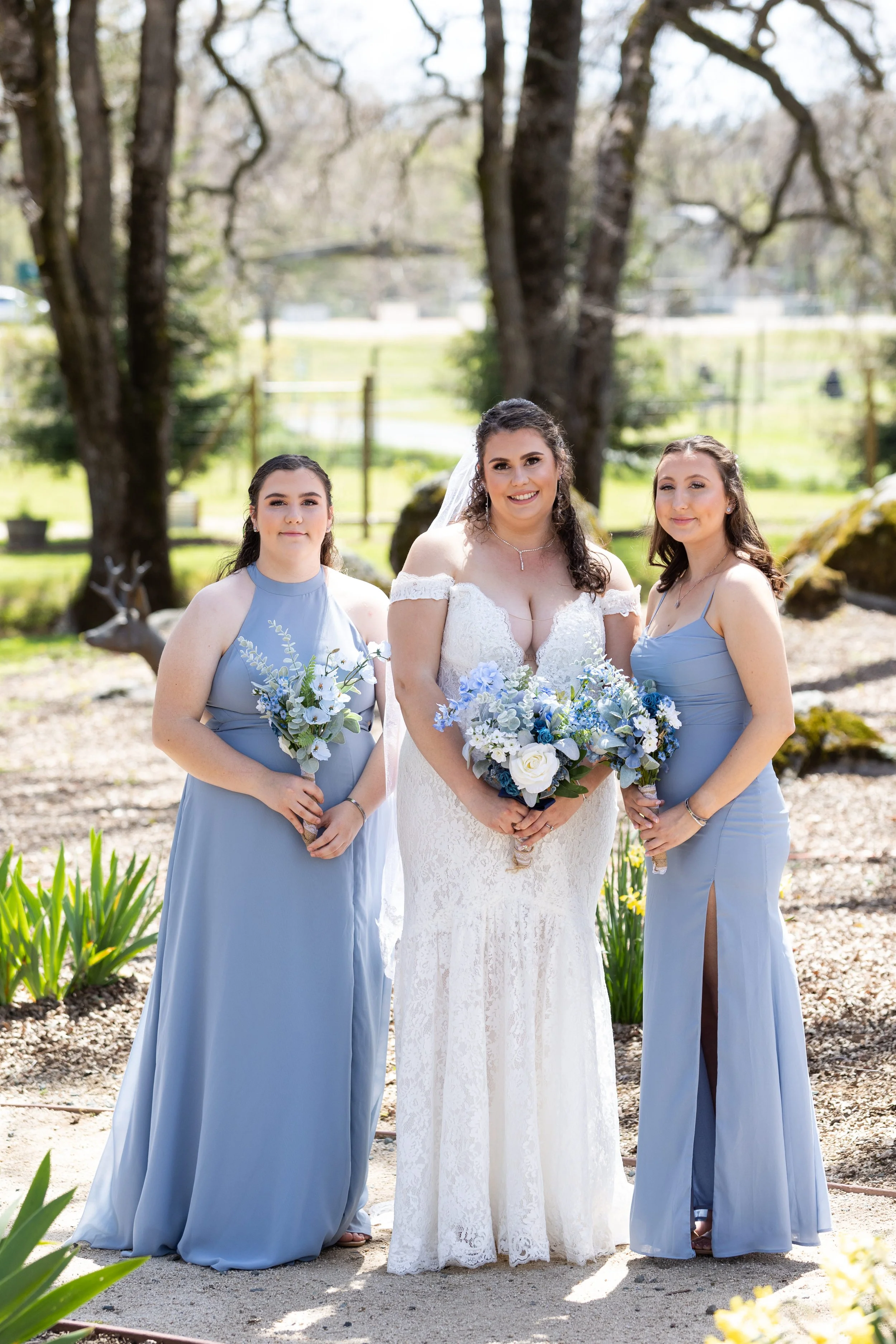 Three women at a wedding outdoors, with the bride in the center wearing a white lace gown holding a bouquet of blue and white flowers, flanked by two bridesmaids in blue dresses holding smaller bouquets. They are standing on a garden path with trees