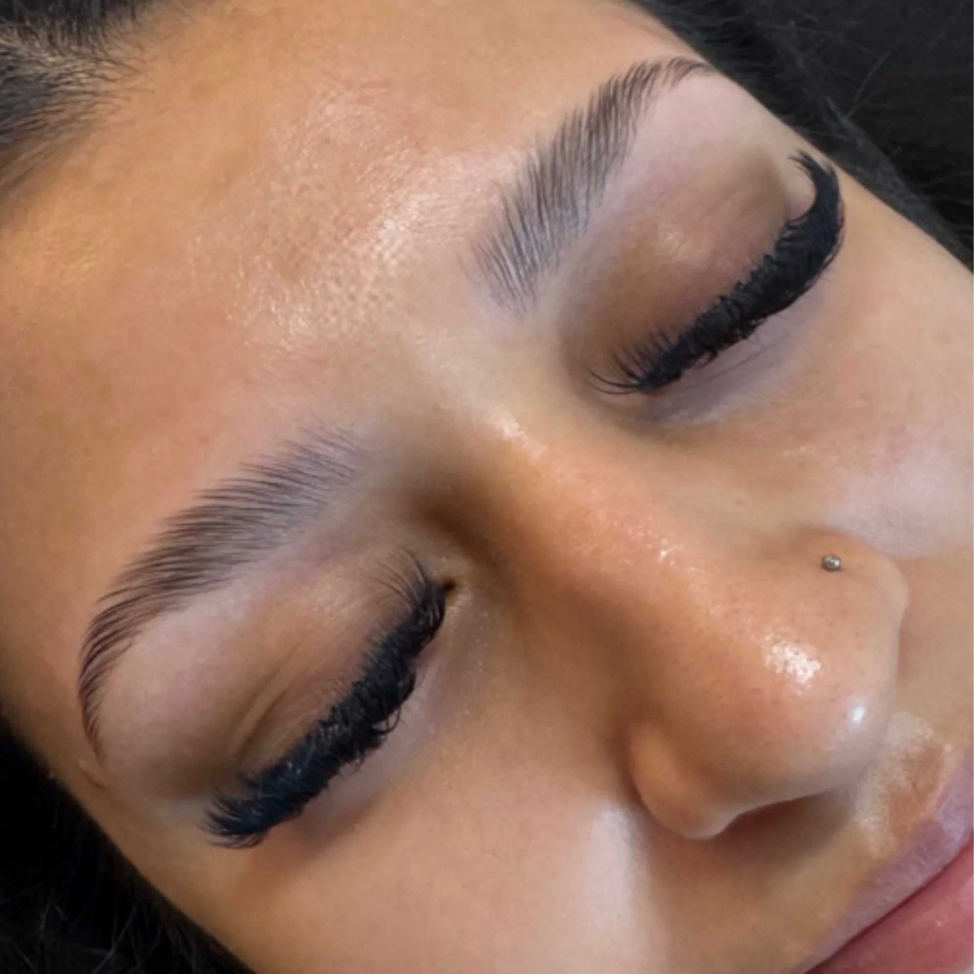 Close-up of a woman's face with closed eyes, showcasing well-groomed eyebrows, long eyeliner, and eyelash extensions. She has a small nose piercing on her nose.