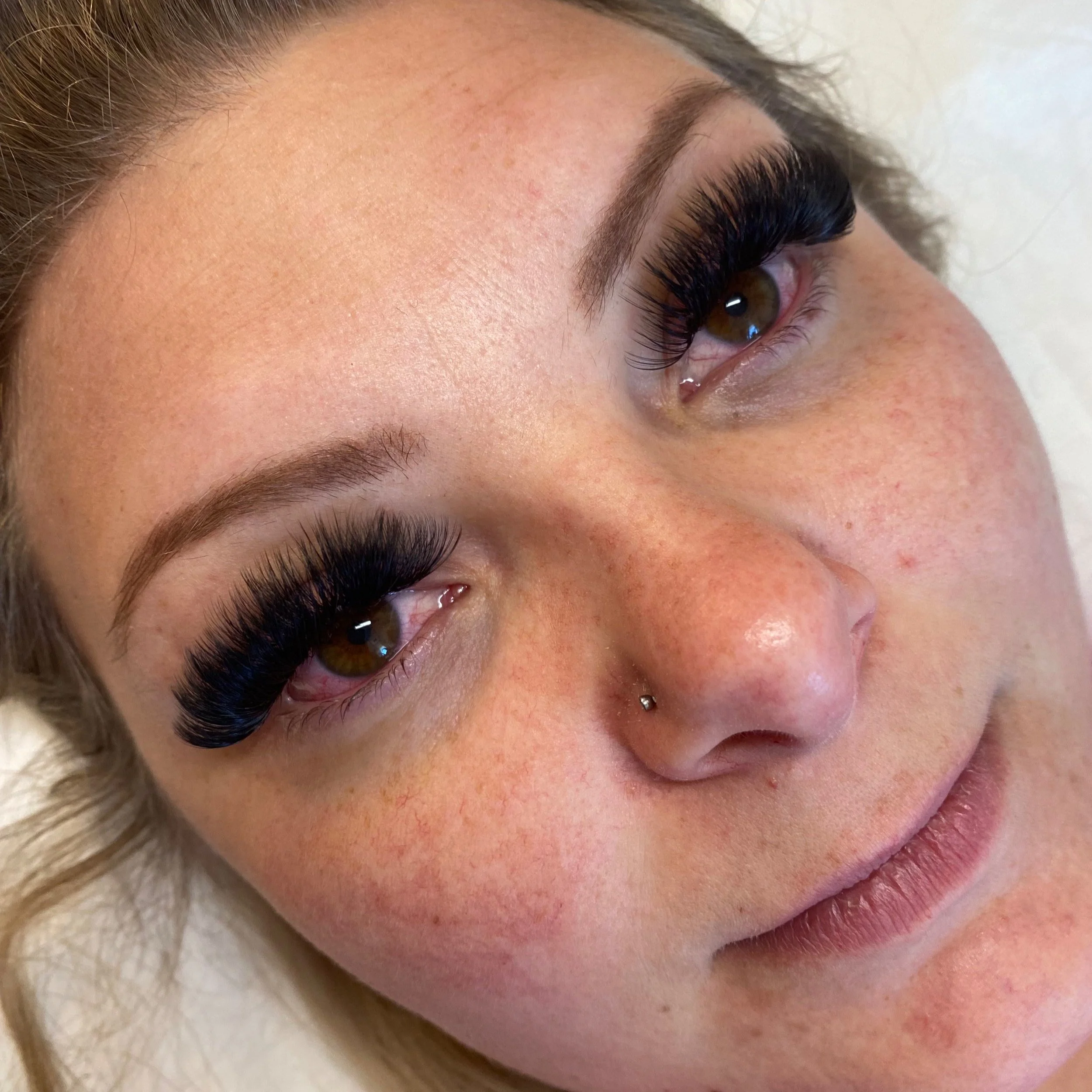 Close-up of a woman's face with eyelash extensions, light skin with freckles, and a nose piercing.