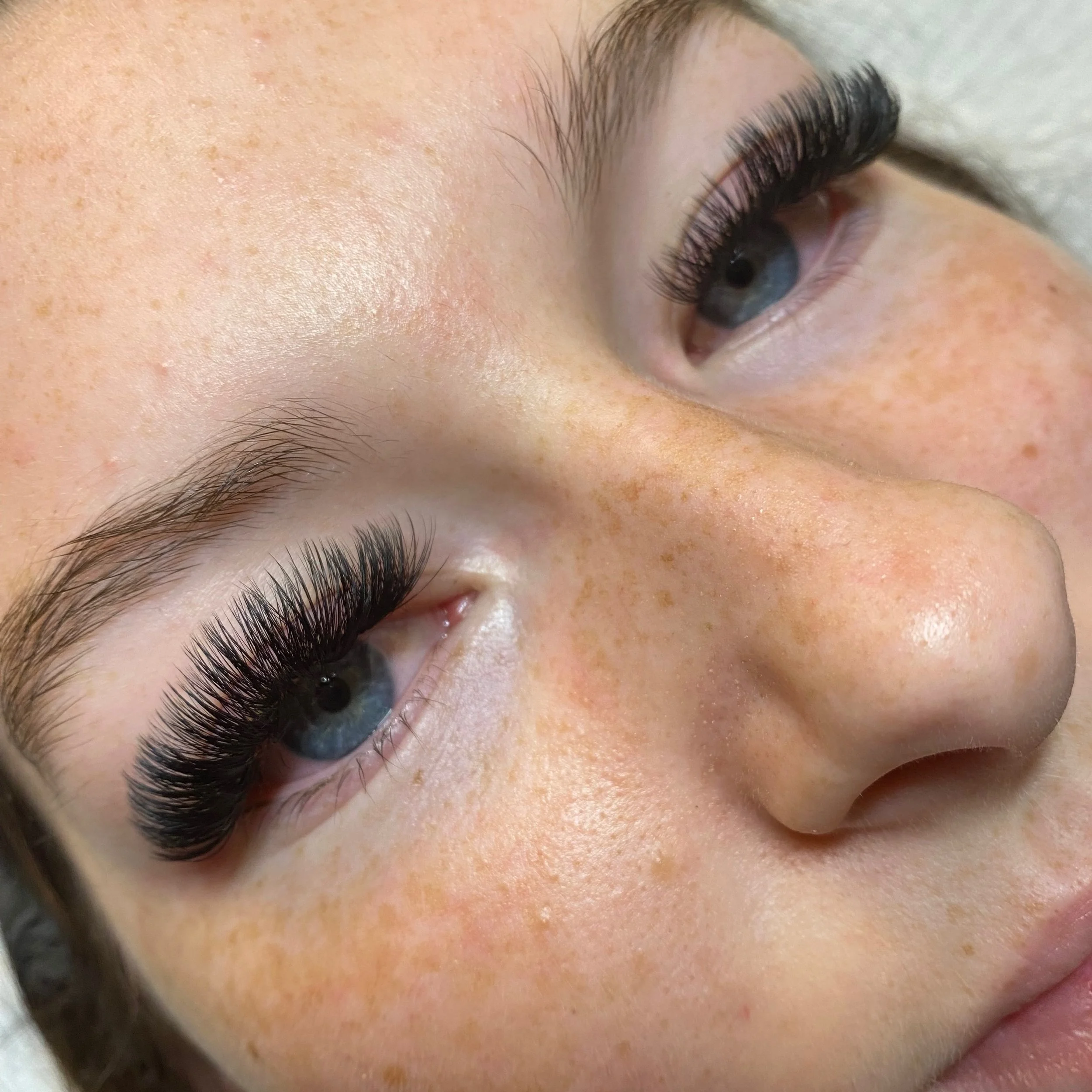 Close-up of a person's face showcasing long, voluminous eyelash extensions with blue eyes and natural freckles.