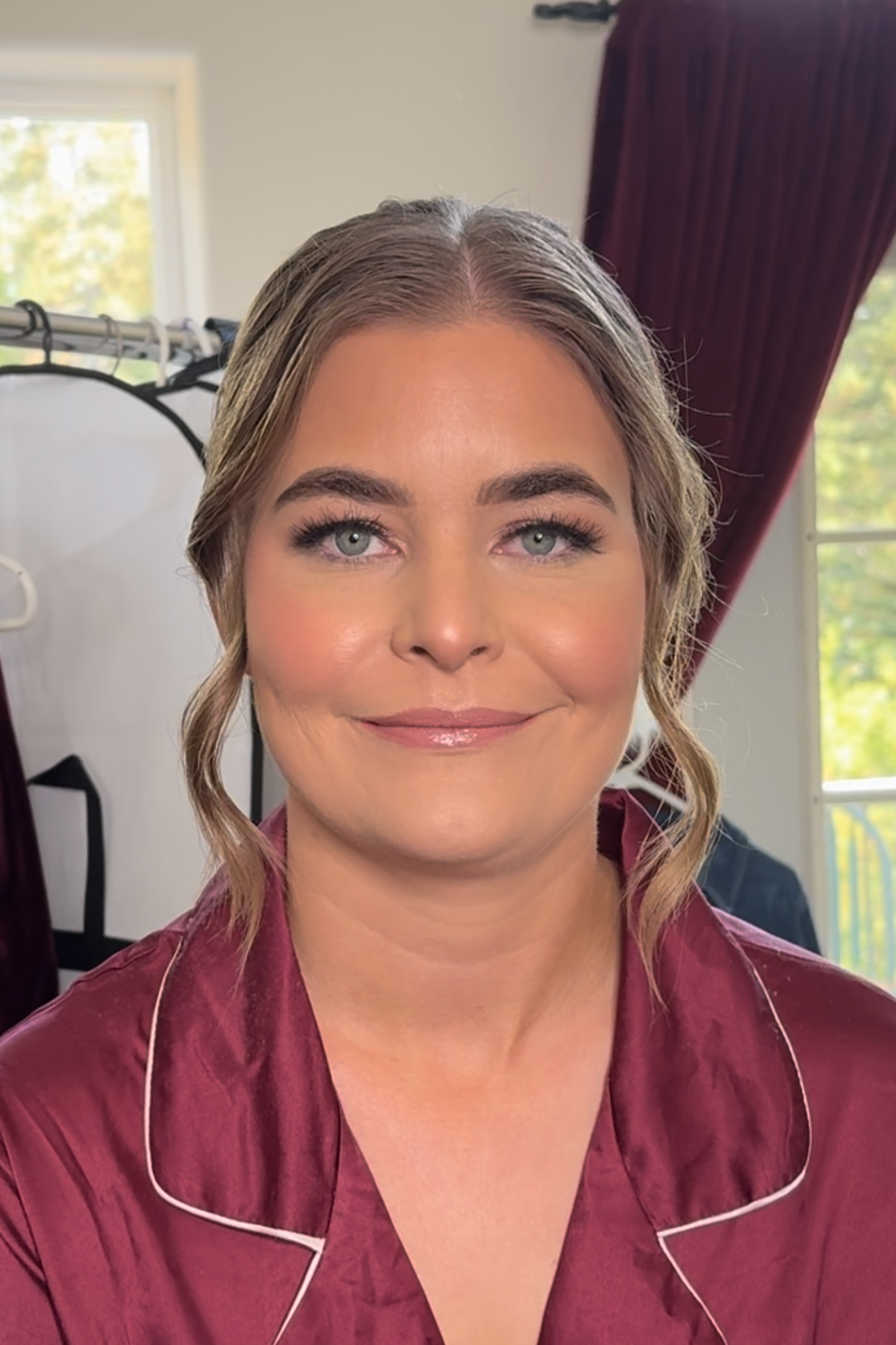 A woman with light skin, blue eyes, wearing makeup, and styled hair, smiling softly in a cozy room with a window showing greenery outside and maroon curtains, wearing a satin burgundy pajama top.