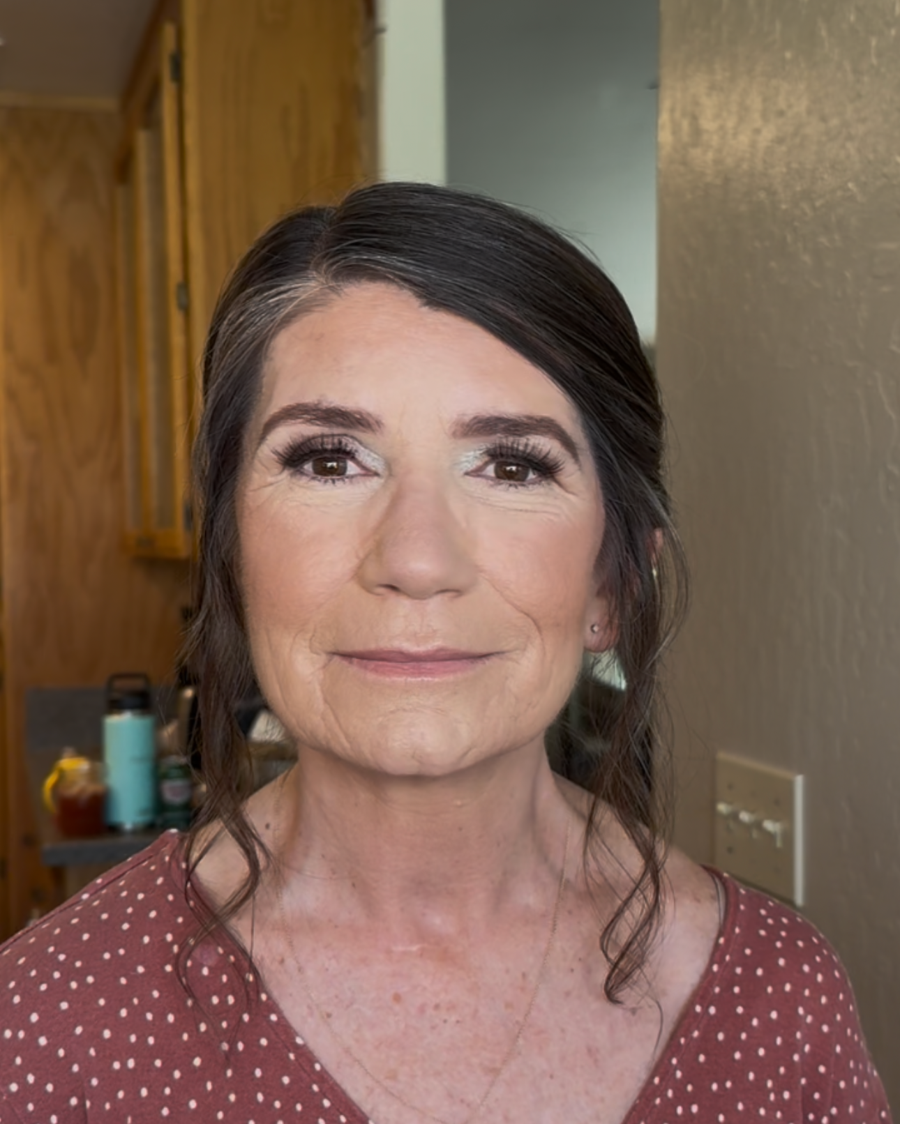A woman with dark hair styled in loose waves, wearing makeup including eyeshadow and mascara, smiling slightly, dressed in a maroon top with white polka dots, standing indoors with wooden cabinets and a countertop with kitchen items in the background