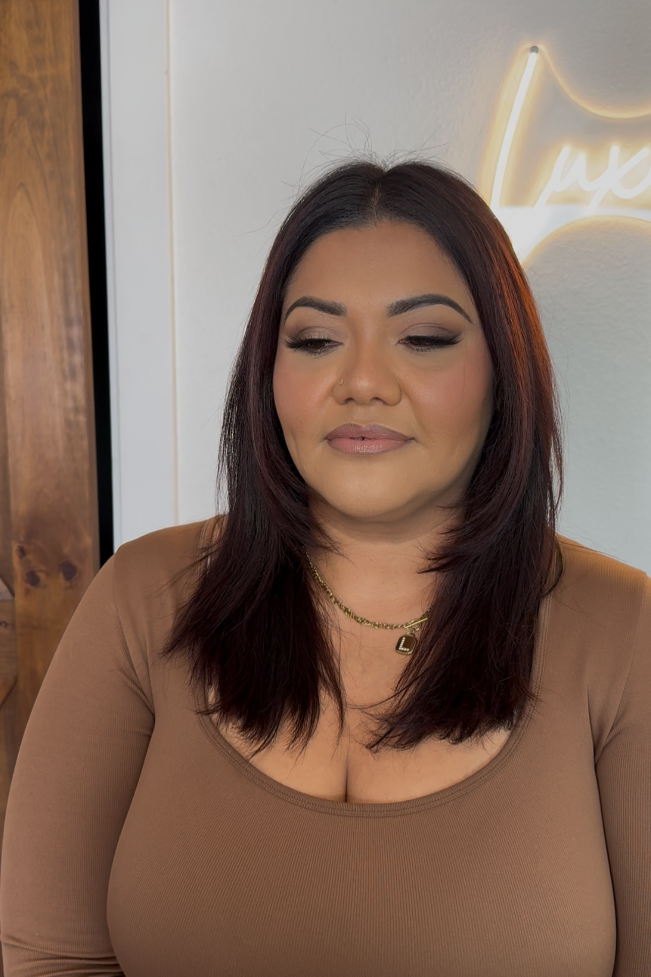 Close-up of a woman with makeup and dark hair, wearing a brown top and a gold necklace, standing inside a room with a white wall and a neon sign in the background.