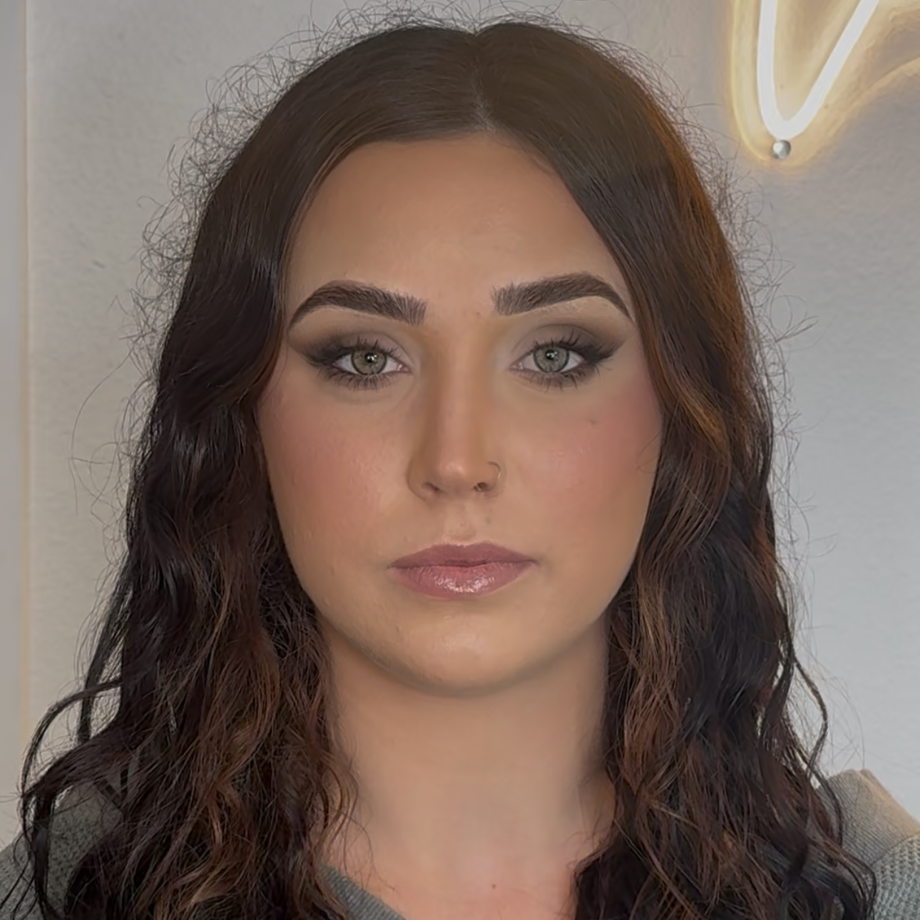 Close-up of a woman with wavy dark brown hair and makeup, including gray eyeshadow, eyeliner, and nude lipstick.