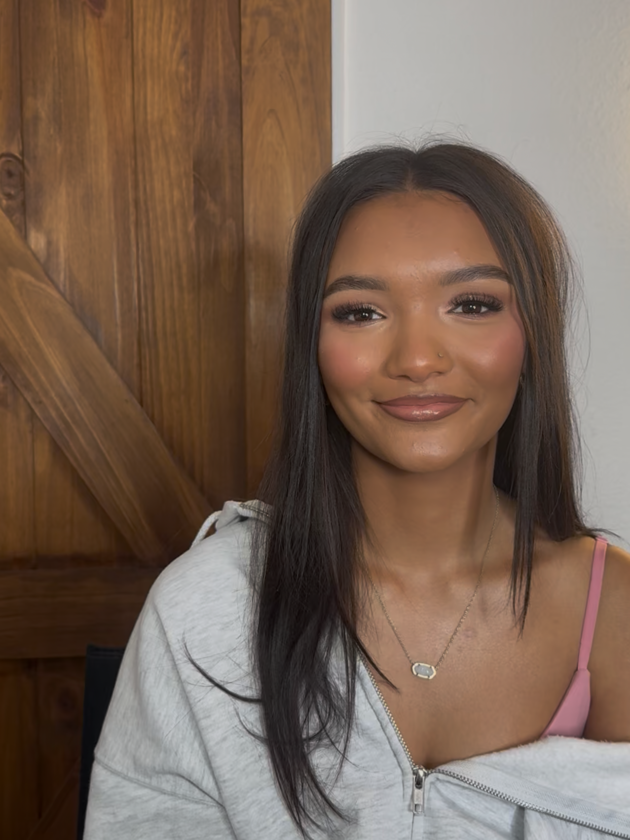 Close-up of a smiling woman with shoulder-length dark hair, wearing a pink tank top, a light gray hoodie partially zipped, a necklace with a pendant, and natural makeup.