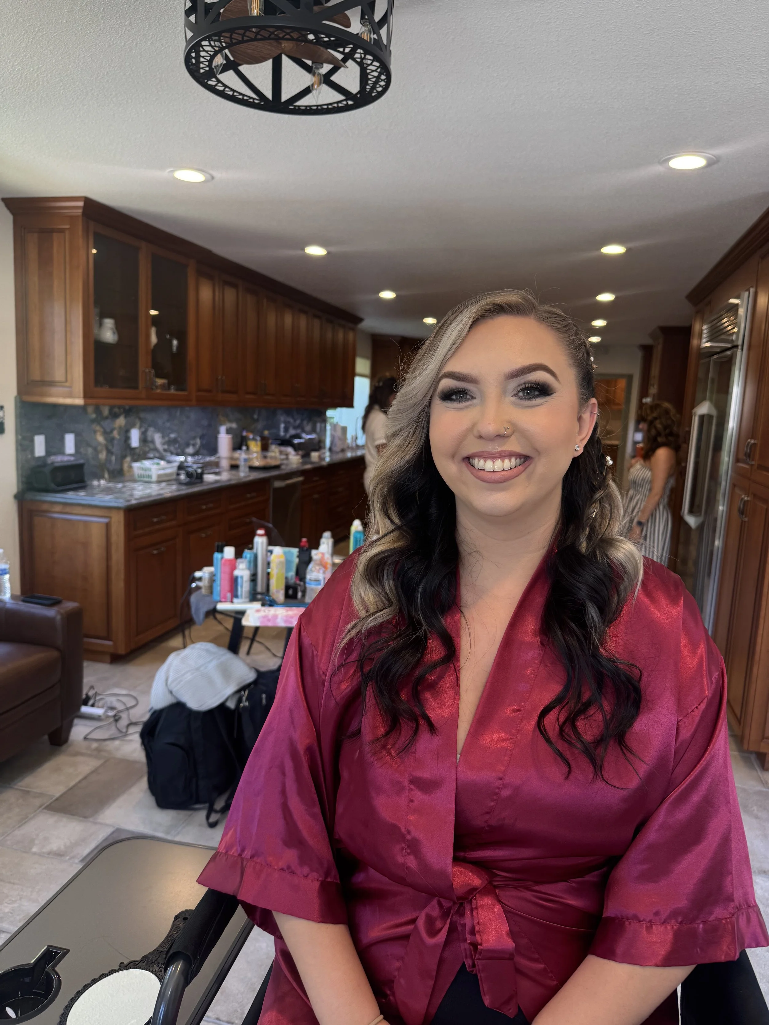 A woman smiling in a pink satin robe in a kitchen setting, with hair and makeup done, likely preparing for an event or photo.