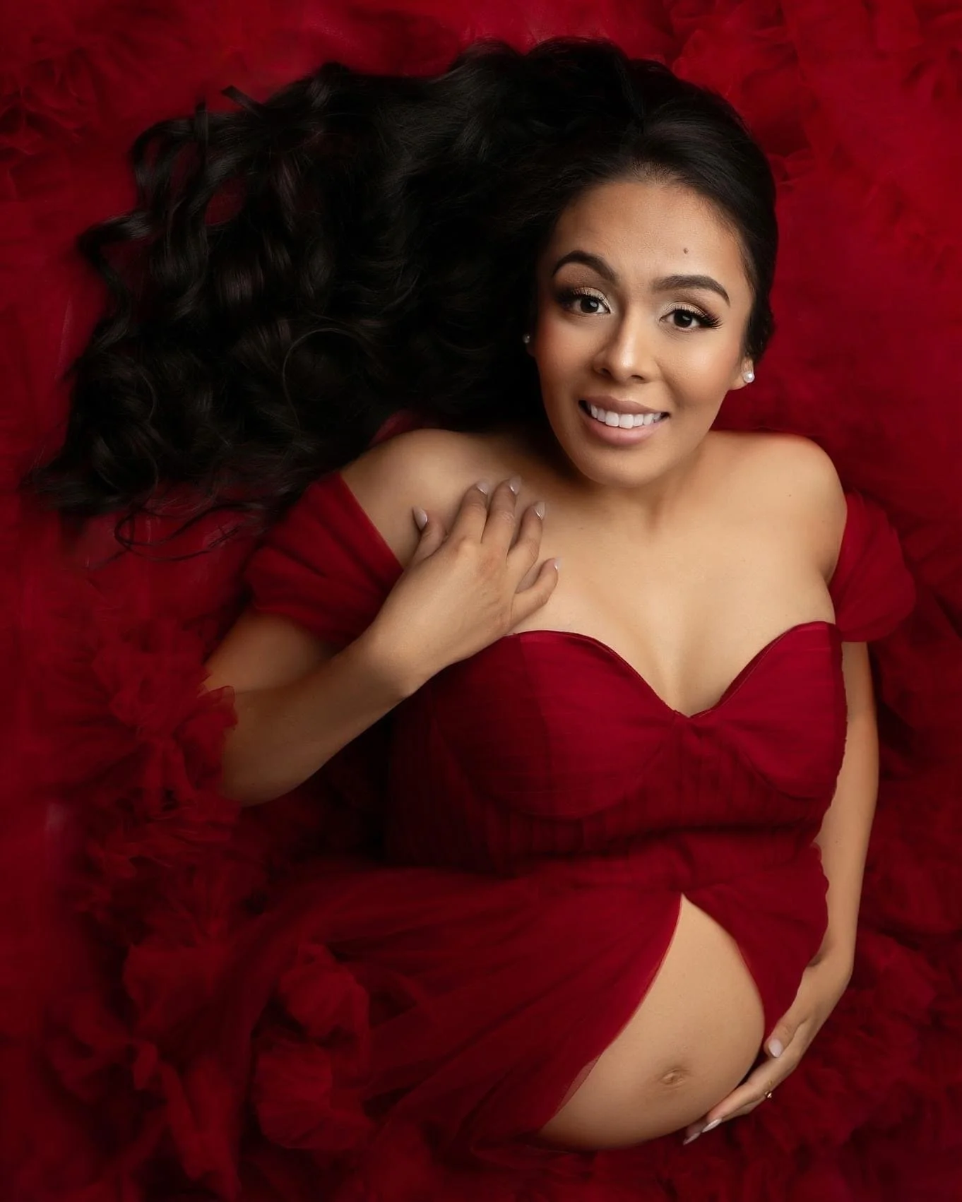 A pregnant woman lying on a red fabric surface, wearing a red dress, smiling, with long dark curly hair.