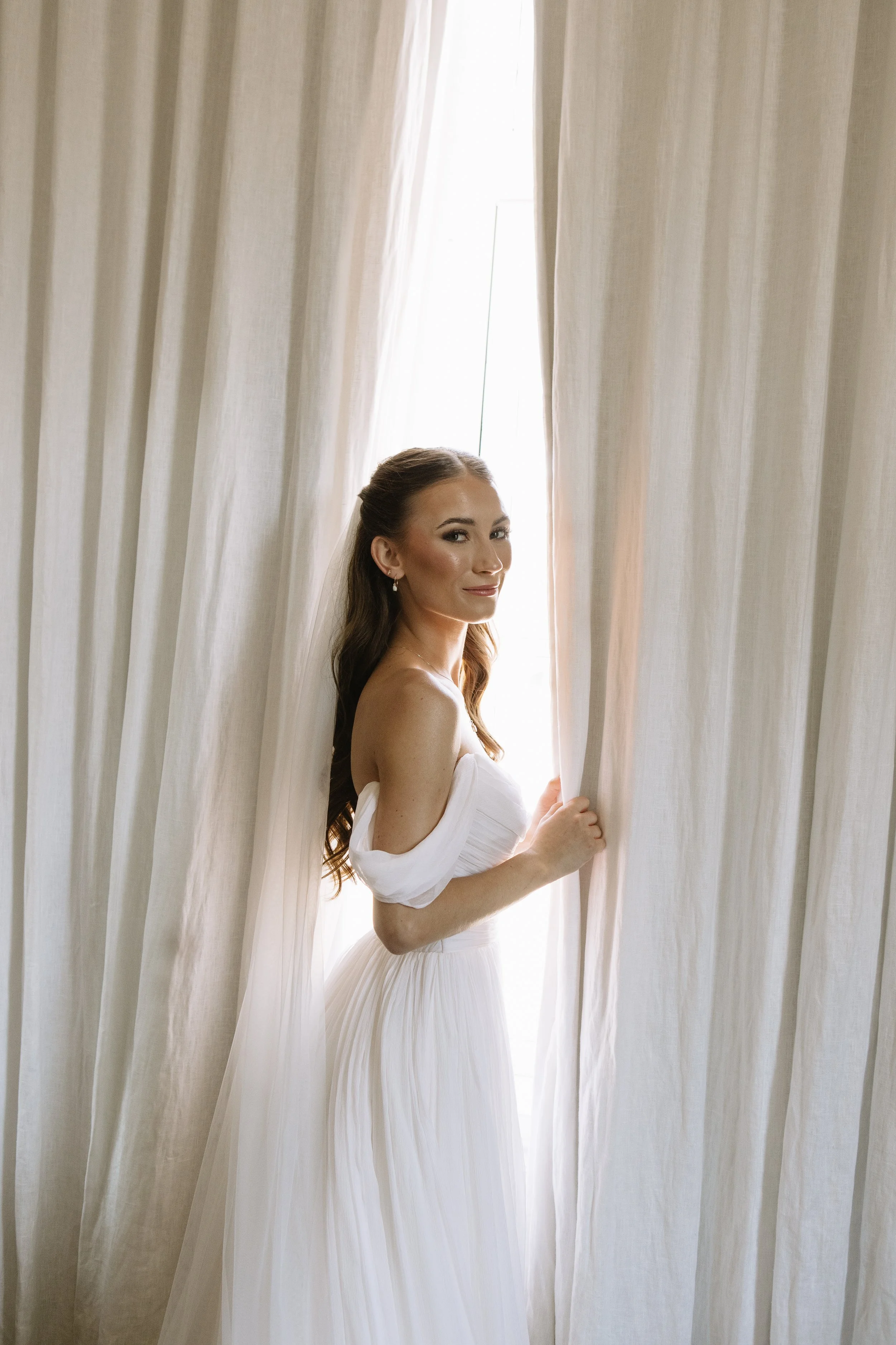 A woman in a white dress standing behind cream-colored curtains, holding the curtains and looking at the camera.