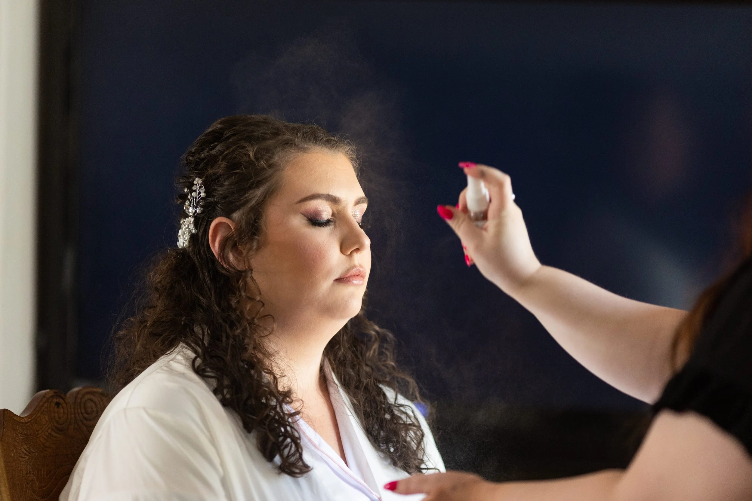 A woman with curly brown hair and a decorative hair clip is having makeup applied by another person who is holding a spray bottle.
