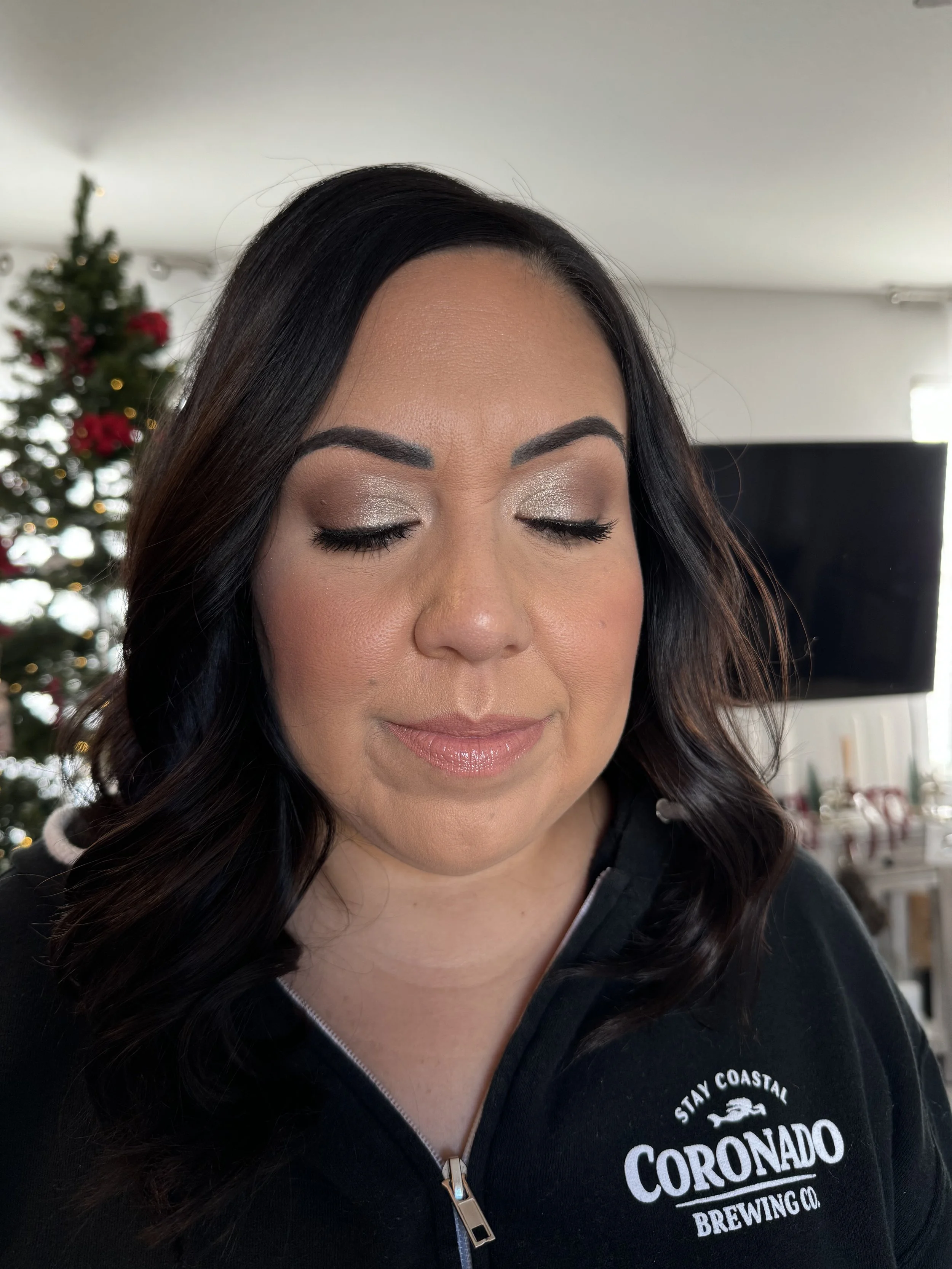 A woman with closed eyes, wearing makeup, and a black zip-up hoodie with the logo of Coronado Brewing Co. in front of a decorated Christmas tree.