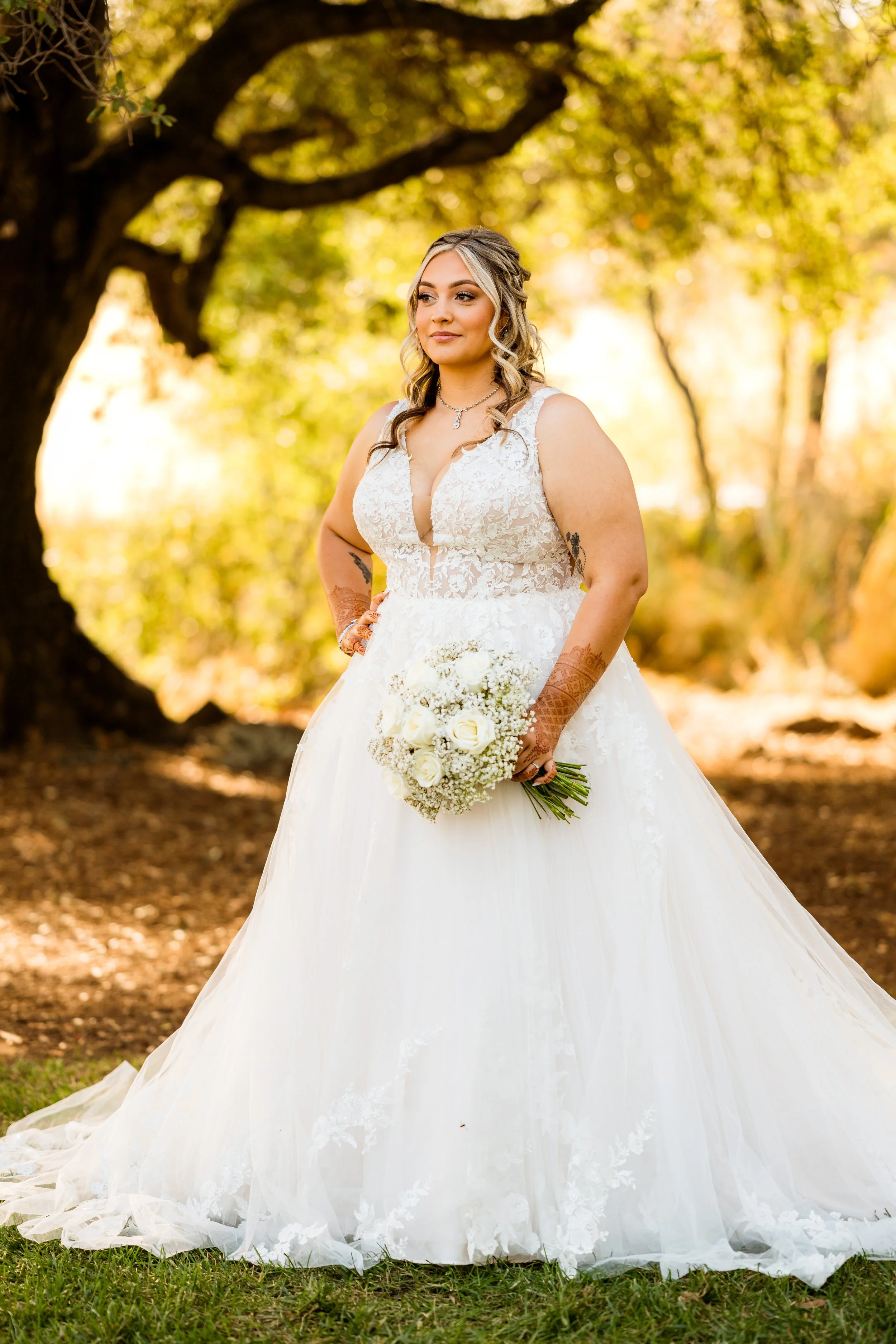 A bride in a white wedding gown holding a bouquet of white roses and baby's breath, standing outdoors in a wooded area with sunlight filtering through trees.