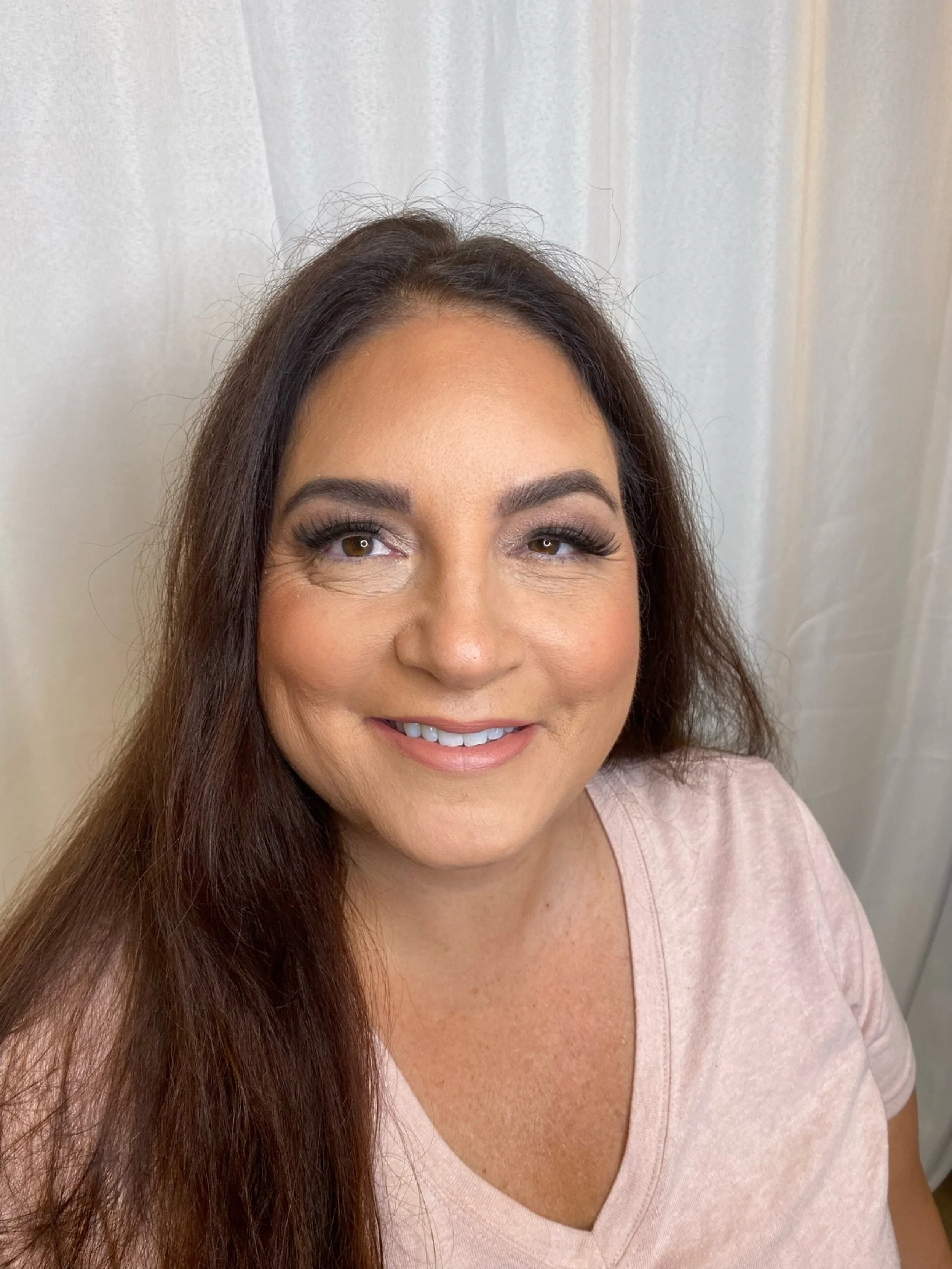 A woman with brown hair and makeup smiling at the camera in front of a light-colored curtain.