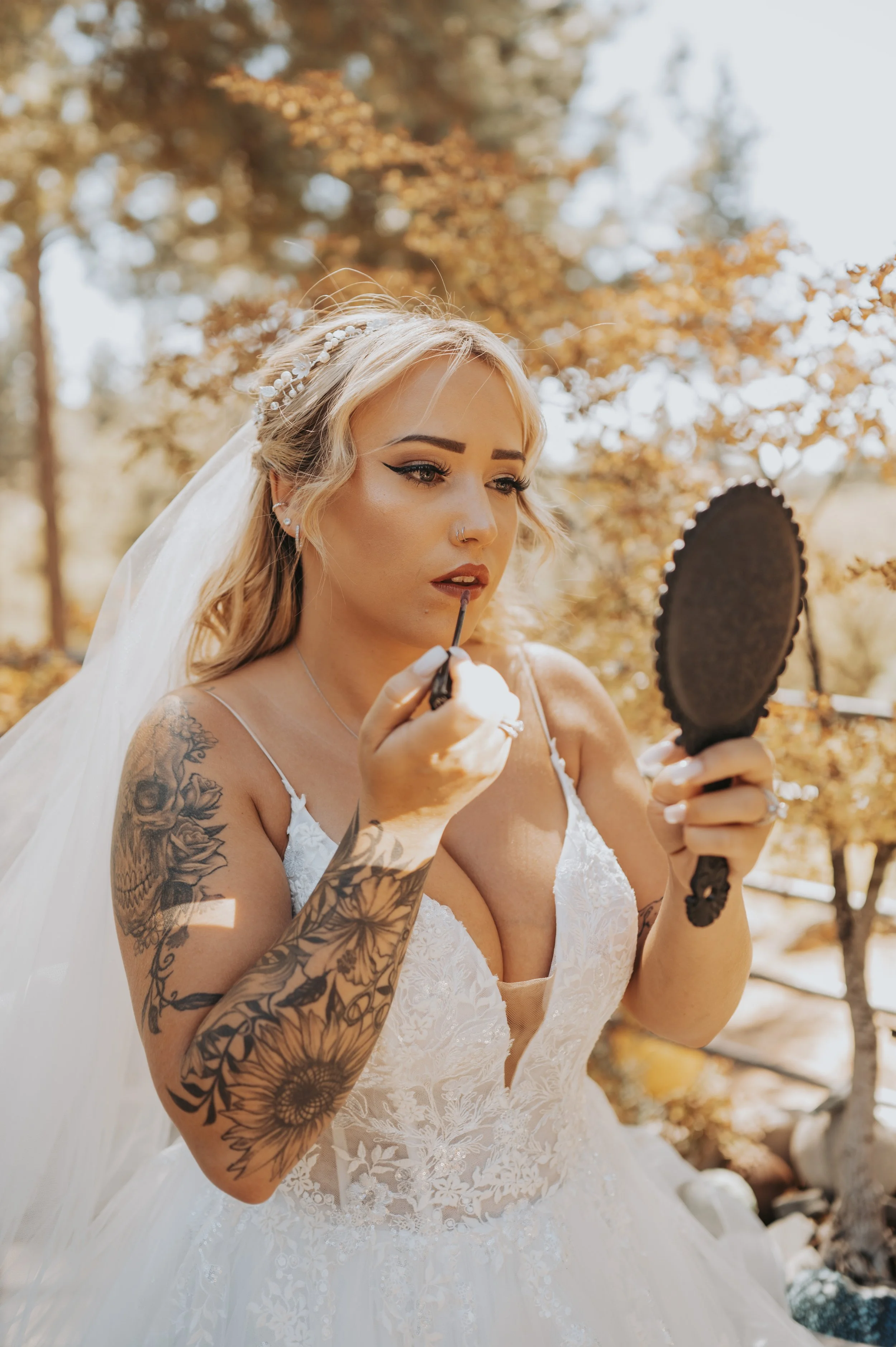 A woman in a white wedding dress with tattoos on her arm is applying lipstick while looking in a hand mirror outdoors with trees in the background.
