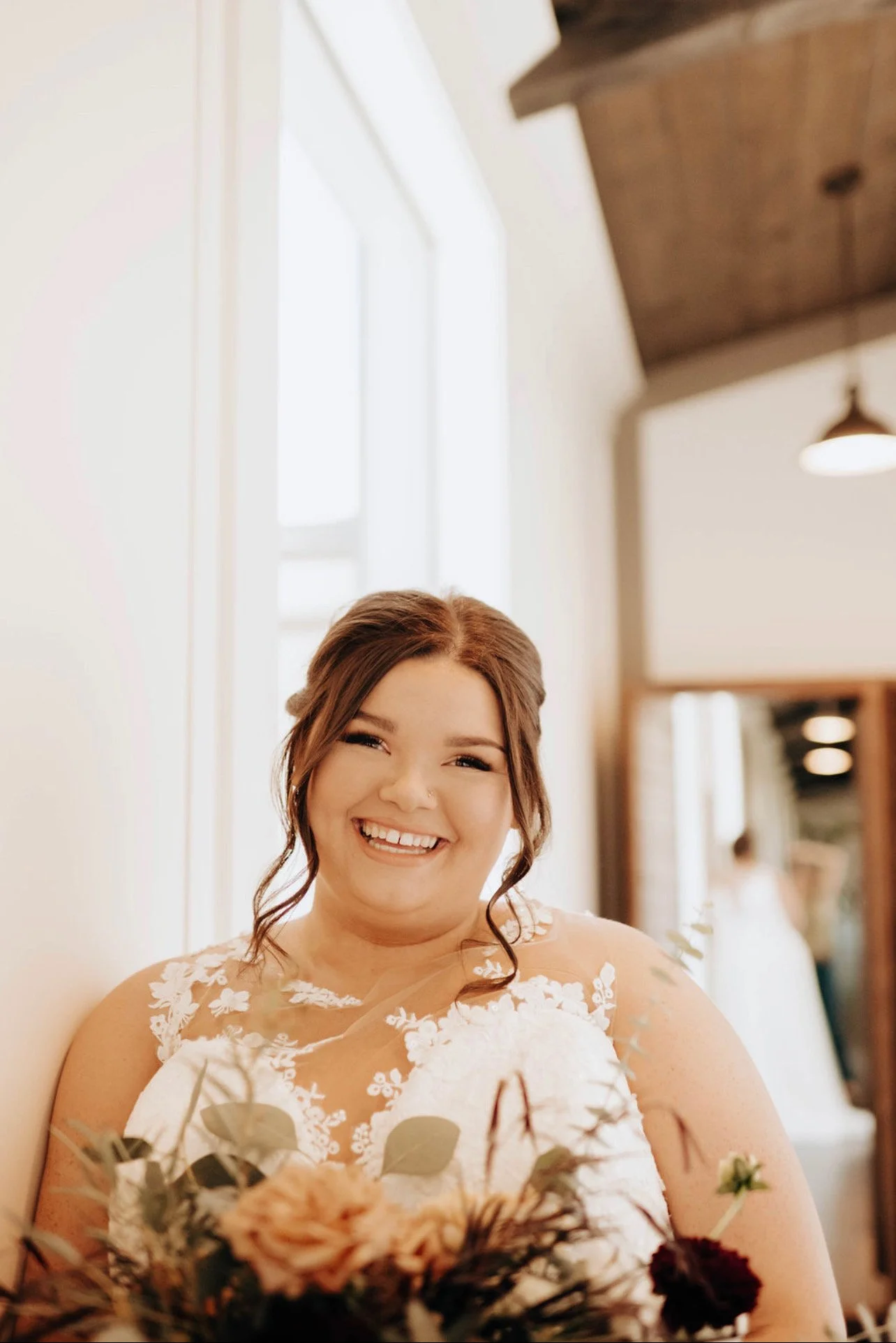 Smiling bride with brown hair and a wedding dress holding a bouquet, in a well-lit room with large windows and wooden ceiling.