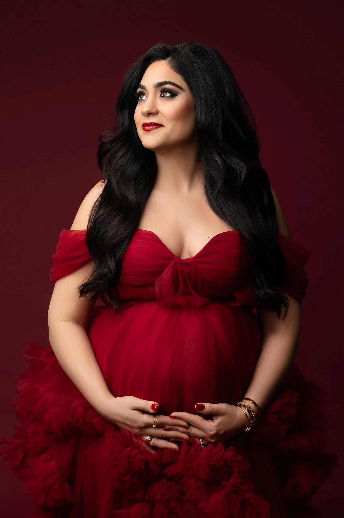 A pregnant woman with long dark hair and makeup, wearing a red off-shoulder dress, holds her belly with both hands against a dark red background.