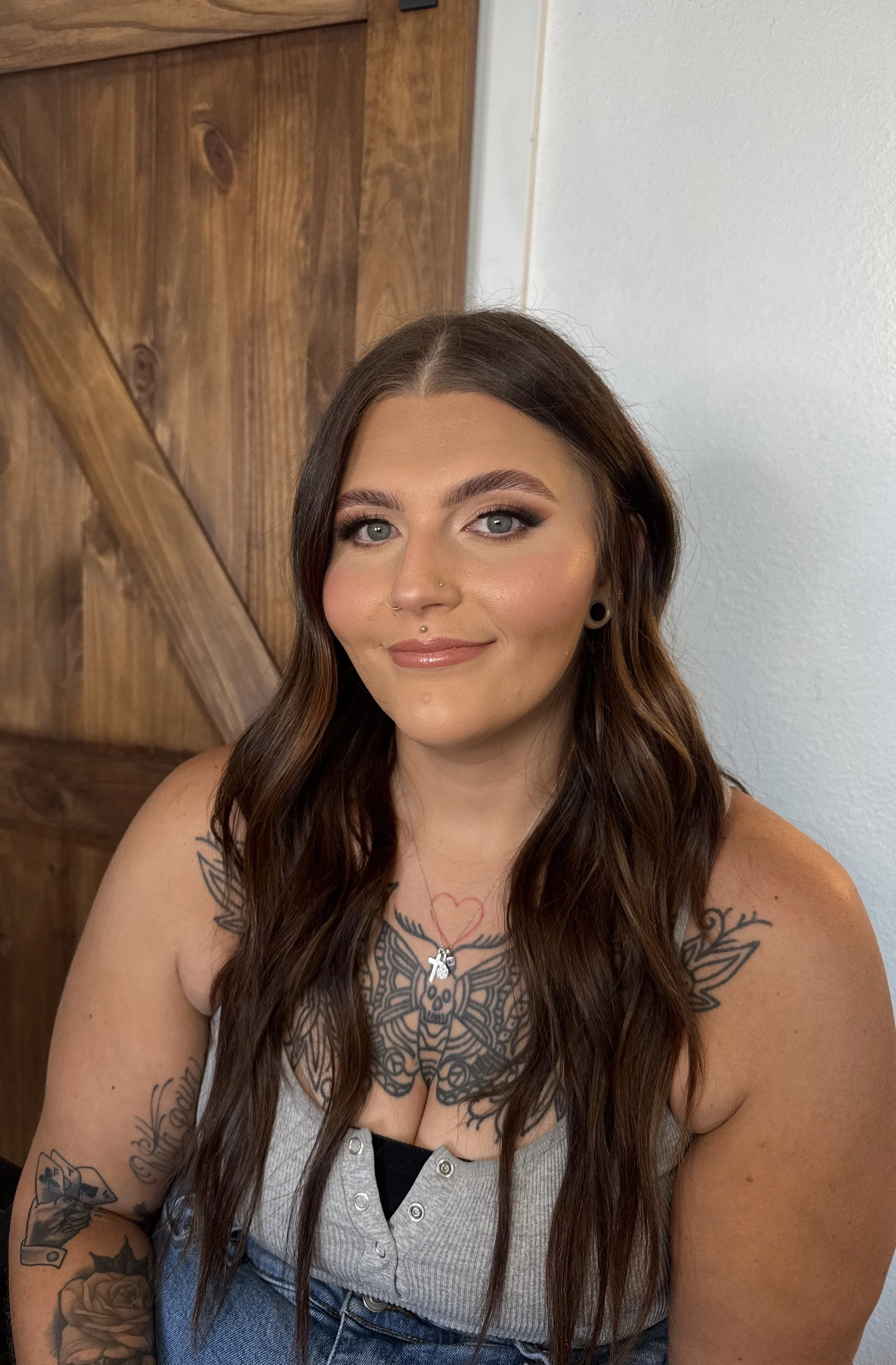 A woman with brown hair and blue eyes smiling, wearing makeup, a gray tank top, and tattoos on her arms and chest, sitting in front of a wooden wall.